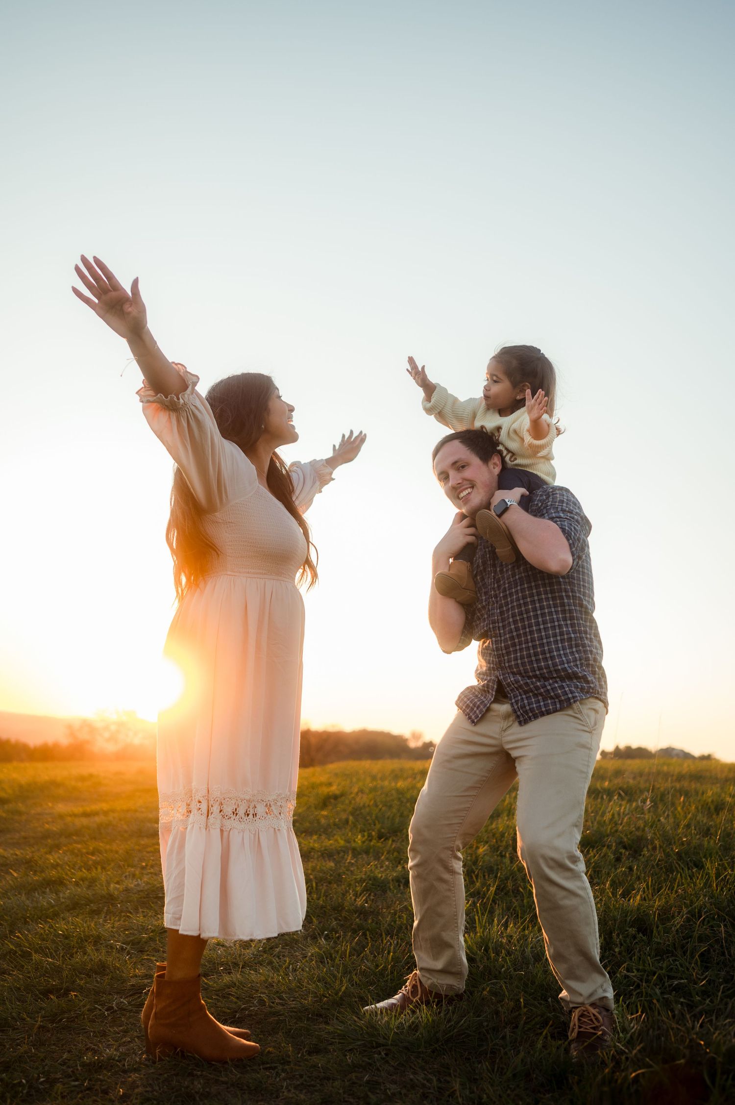 Joyful family at sunset with dad carrying toddler on shoulders and pregnant mom raising arms.