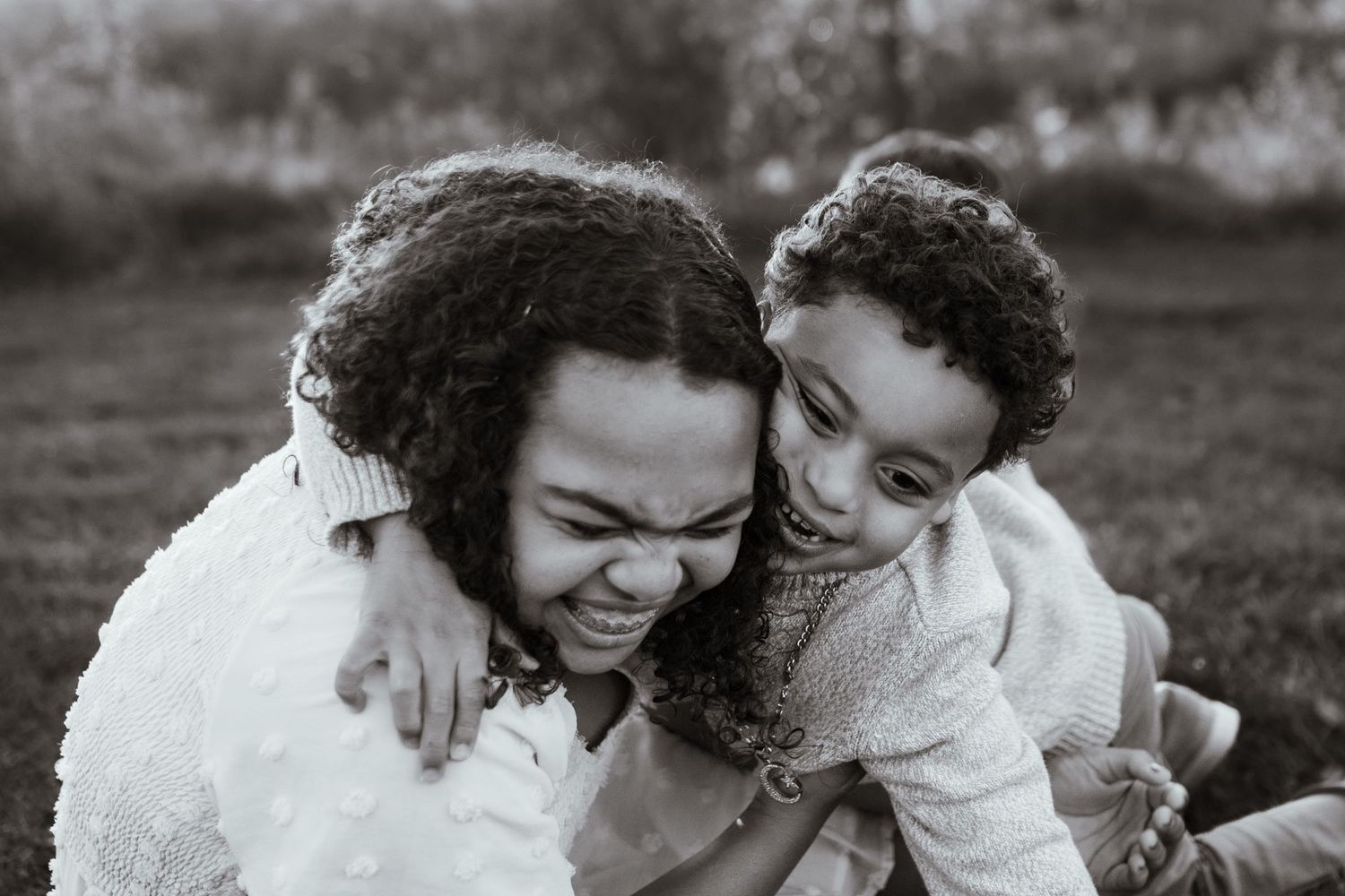 Black and white photo of older sibling laughing while younger child hugs them on grass.