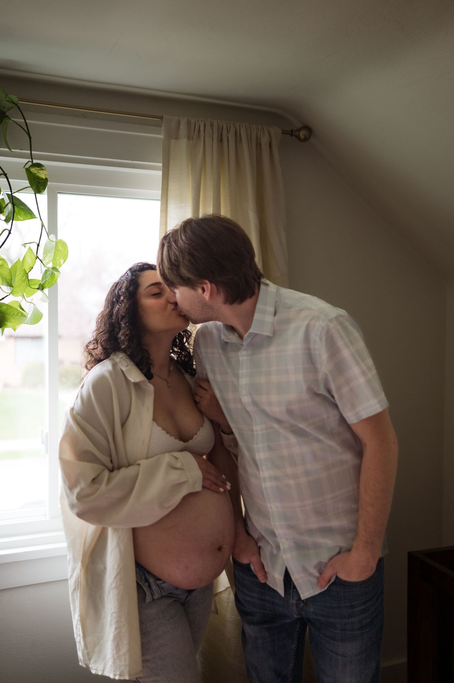 A romantic moment between a couple near a window, with one person wearing white and showing a pregnant belly.