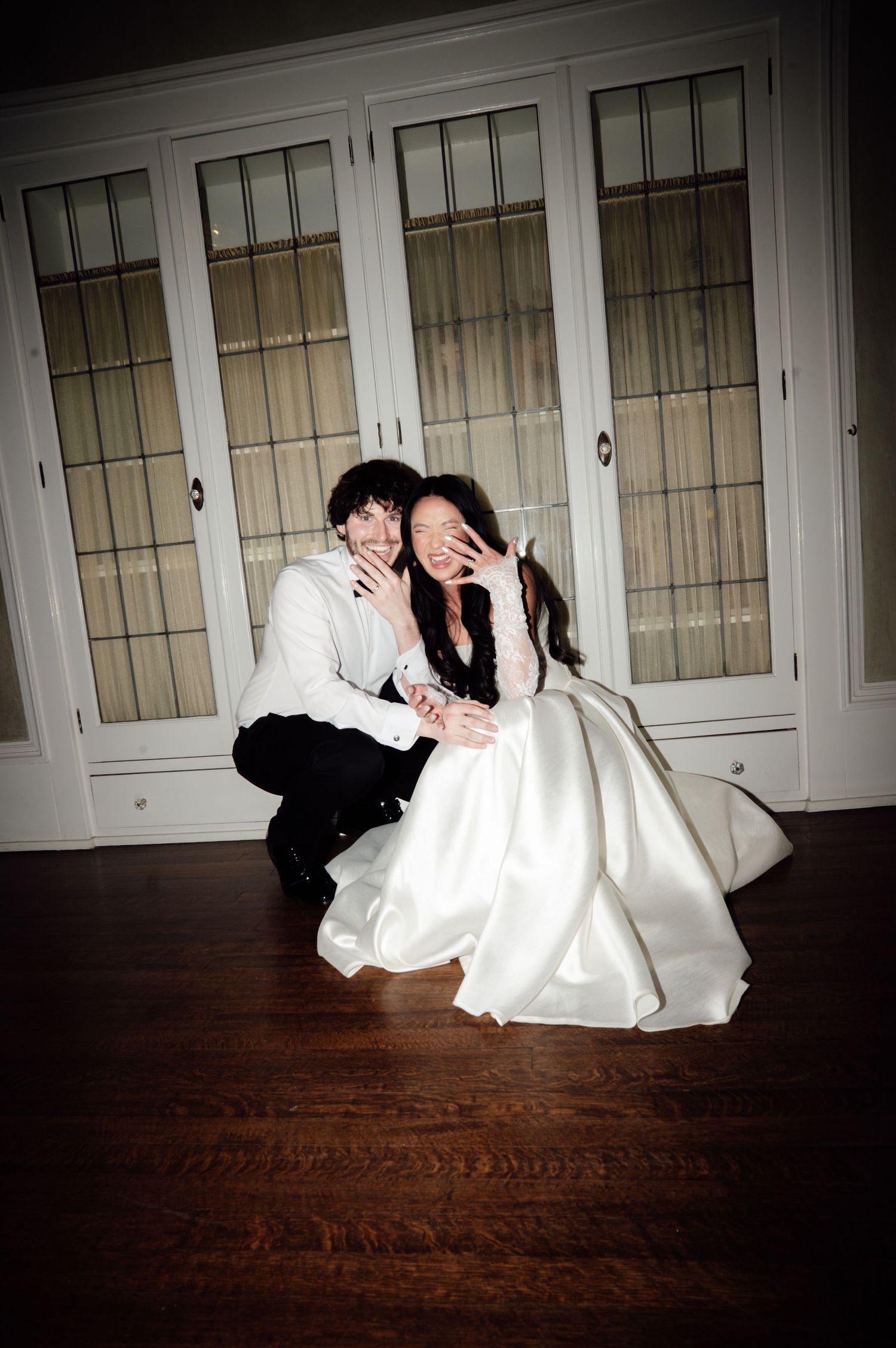 Bride in white gown and groom in black suit crouch on hardwood floor, laughing and covering faces, in front of white cabinets.
