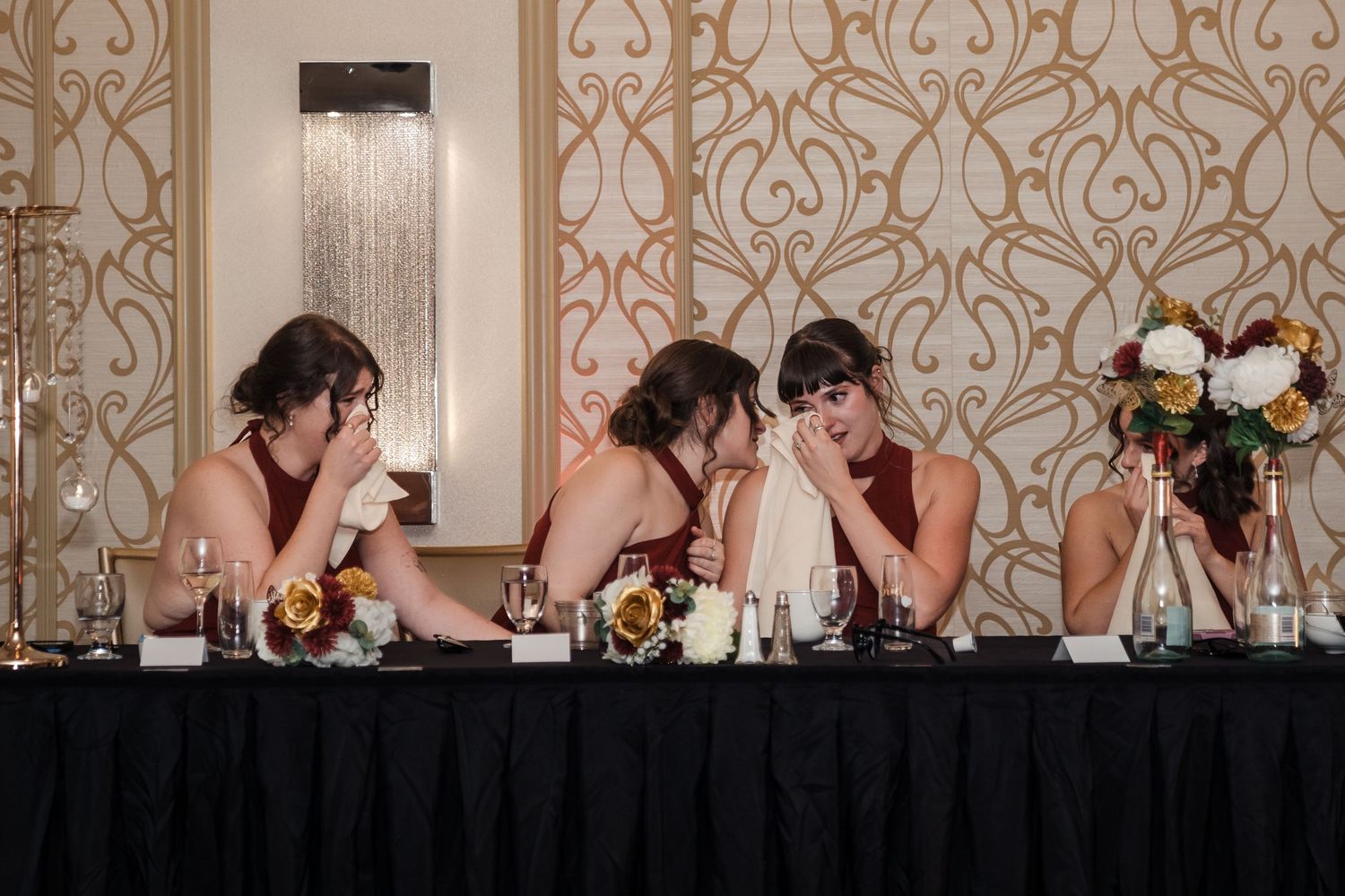 Wedding party members at reception table cover their faces in synchronized playful pose at elegant venue.