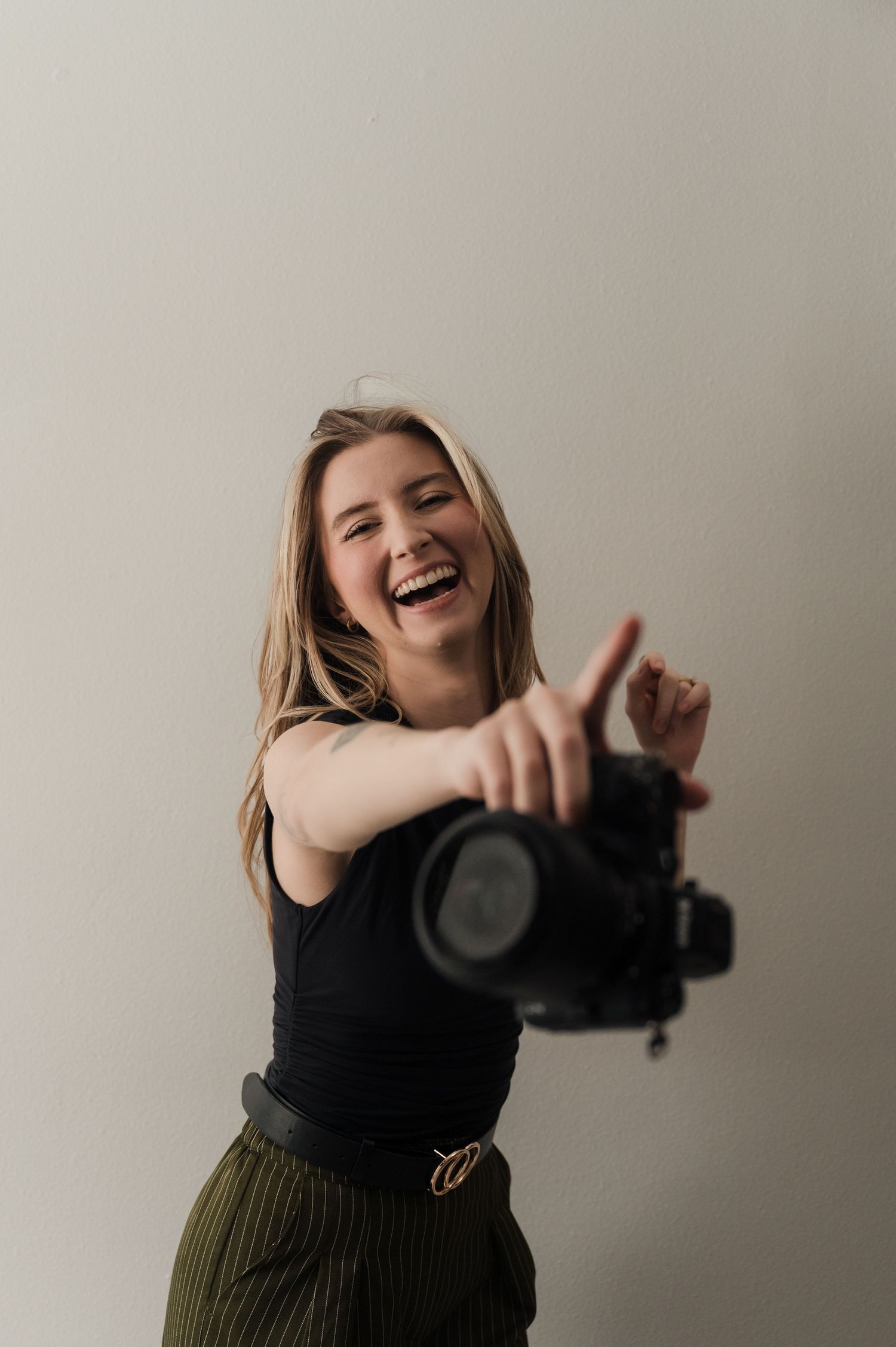 Smiling female photographer laughing while pointing DSLR camera toward viewer against white background.