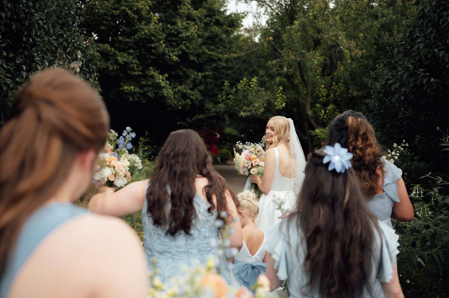Bride in white gown holding bouquet faces bridesmaids in blue dresses in garden setting.