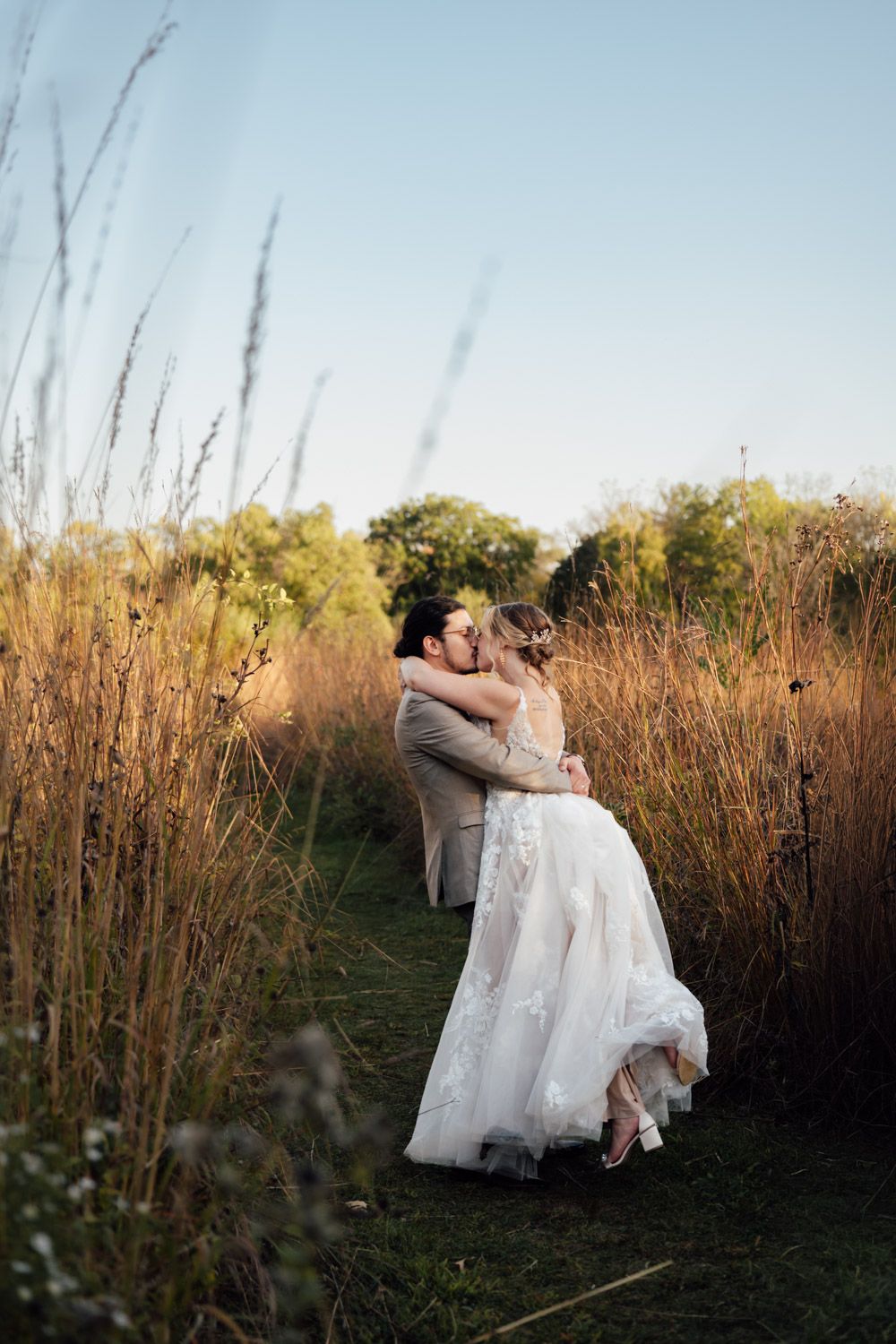 Bride and groom sharing romantic kiss surrounded by tall golden grass at sunset.