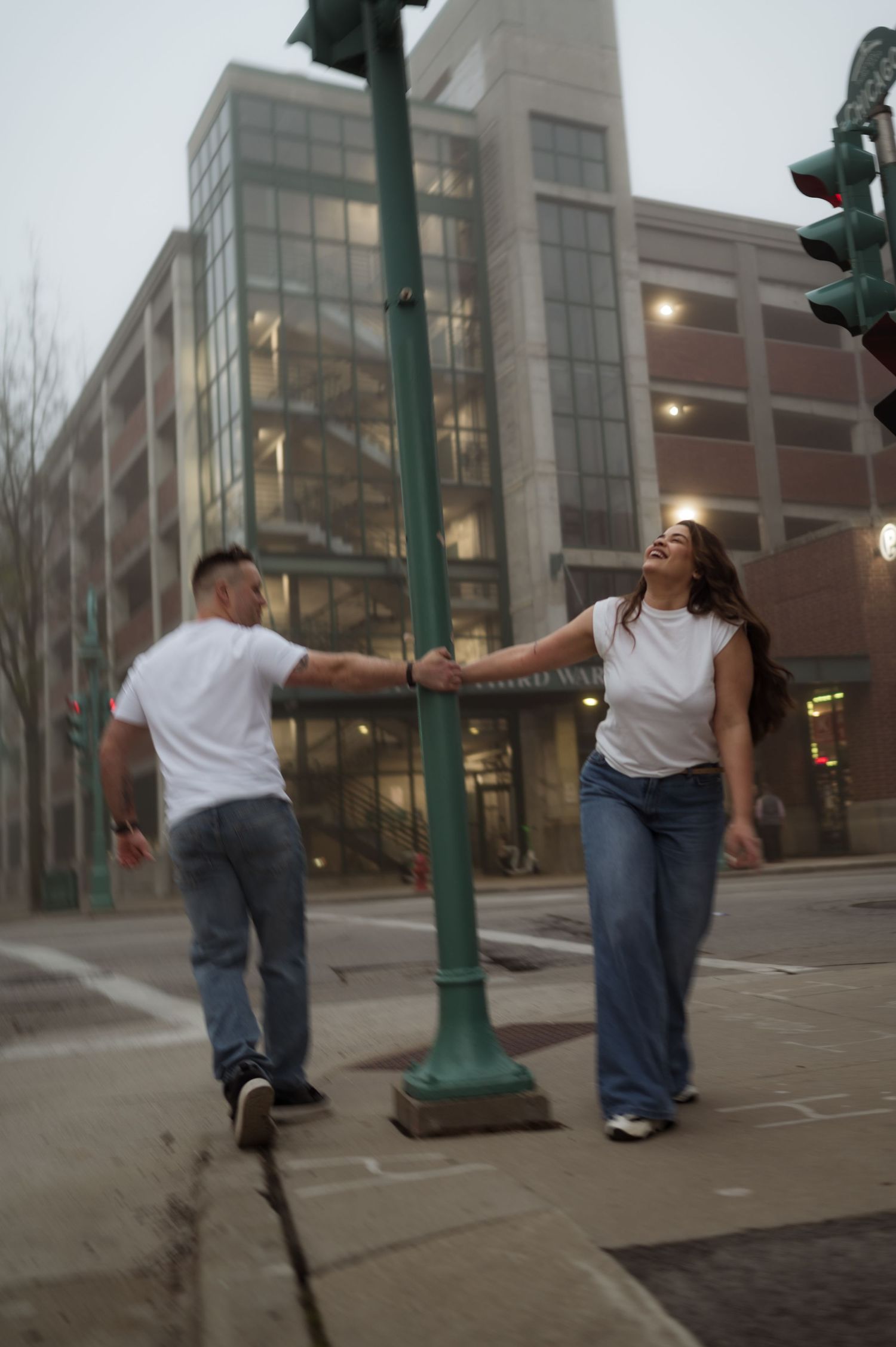 Joyful couple in white tees holding hands while walking on a foggy urban street at dusk.