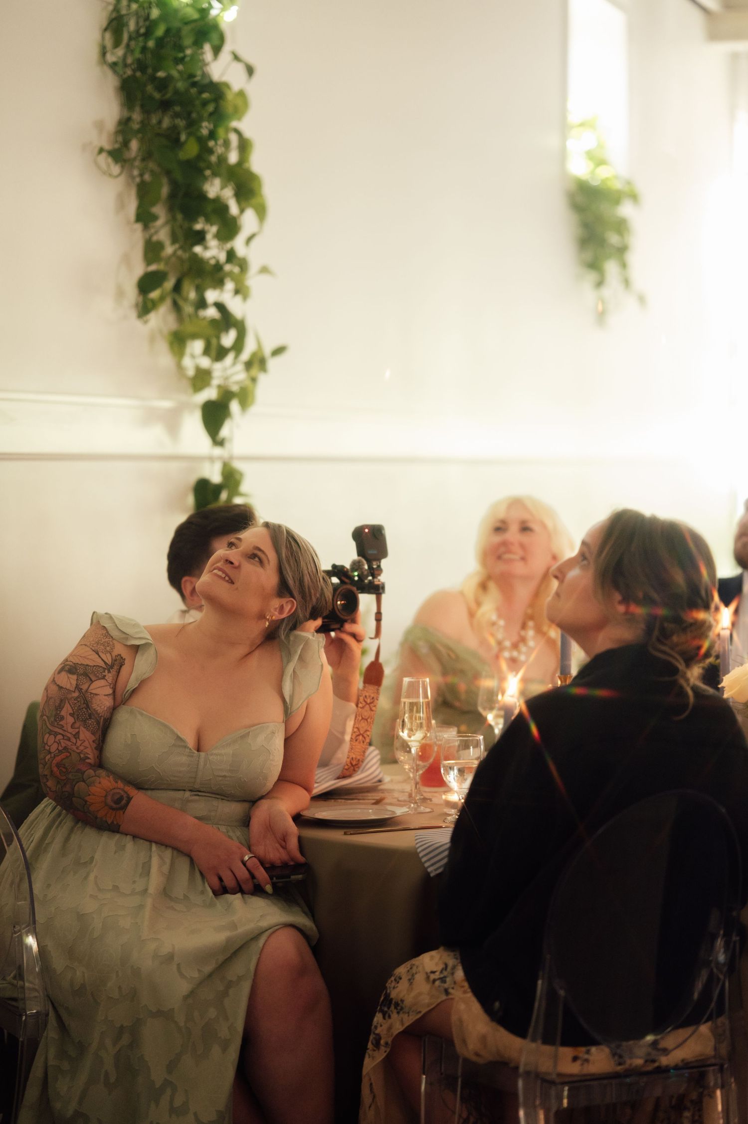 Women in formal attire laugh and socialize at an elegant wedding reception table with champagne glasses and greenery.