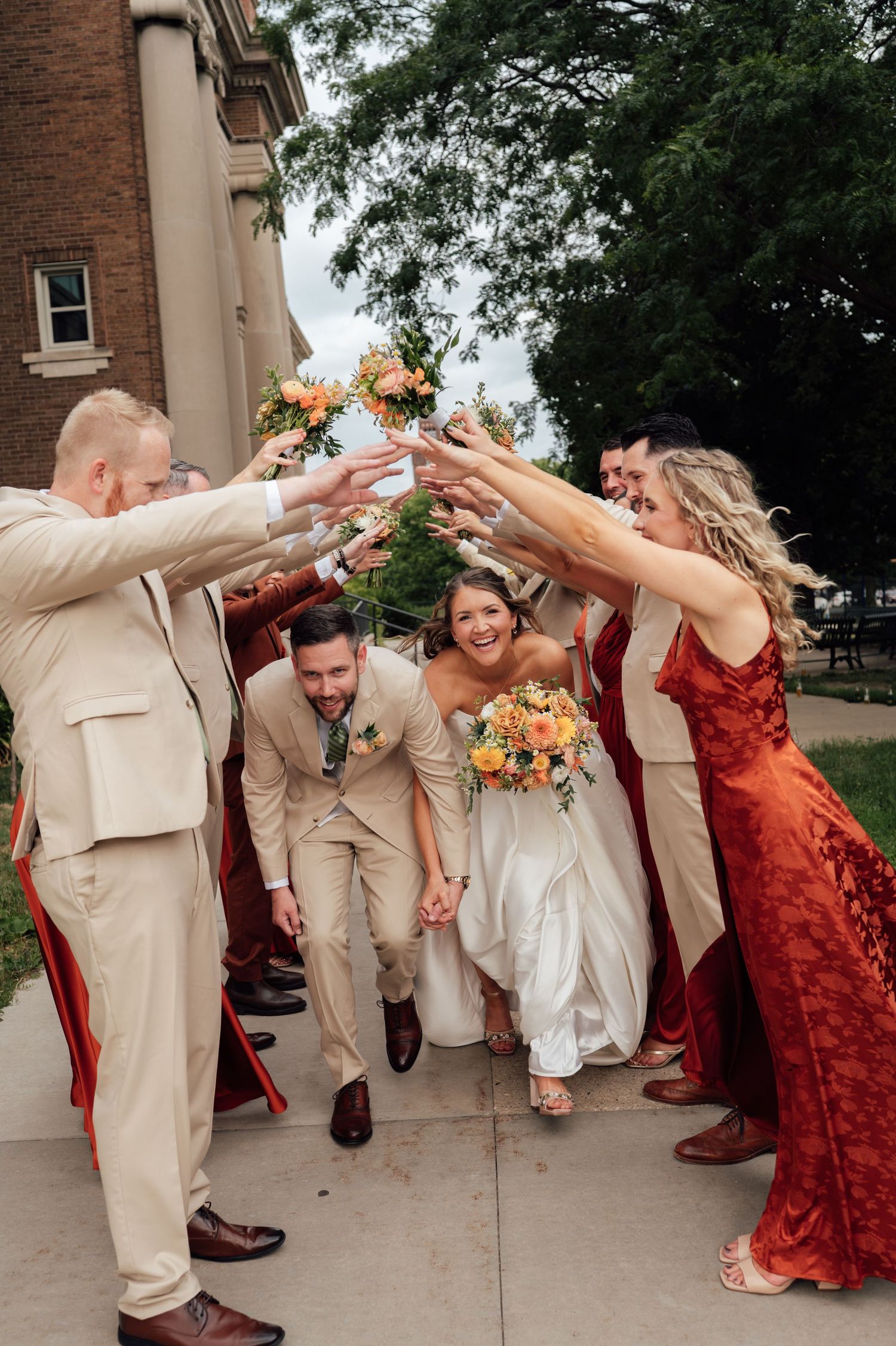 Bride and groom walk through a tunnel of raised hands formed by wedding party in tan and rust red attire outdoors.