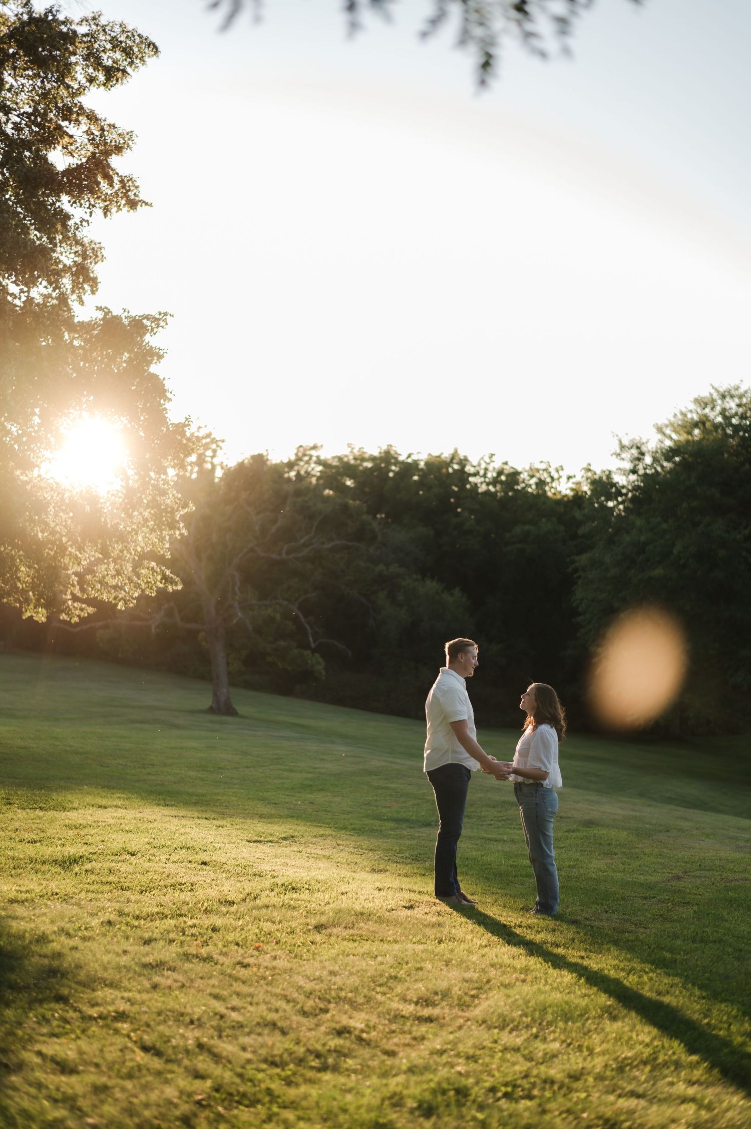 Couple holds hands in a sunlit park at golden hour with lush green trees in the background.