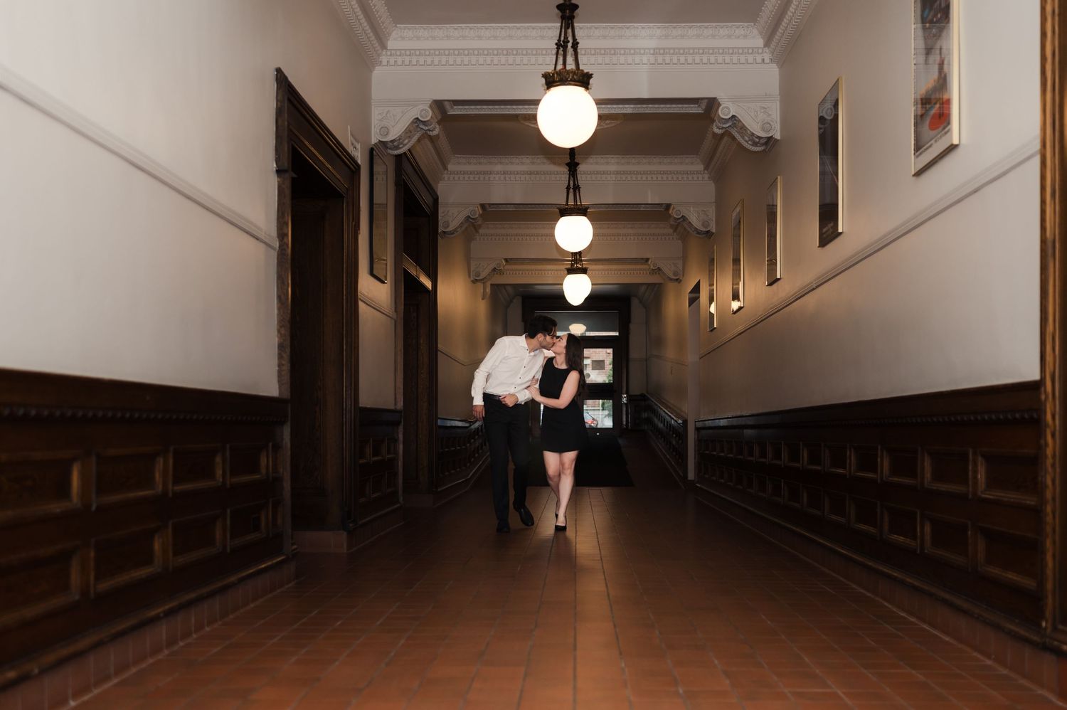 Couple walking hand in hand down an elegant hallway with ornate pendant lights and dark wood paneling.