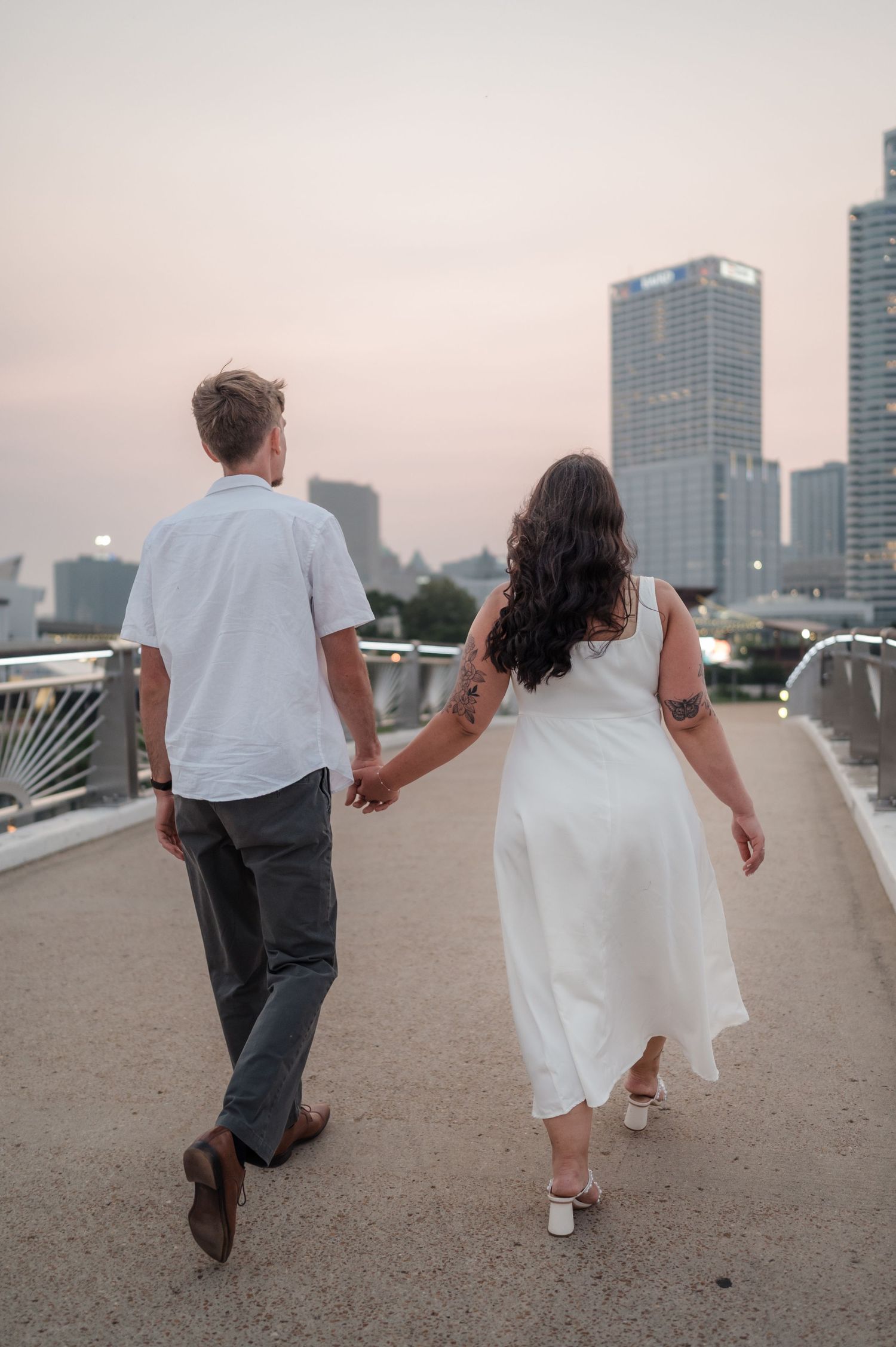 Engaged couple walking hand-in-hand on a bridge at sunset with the Milwaukee skyline behind them.