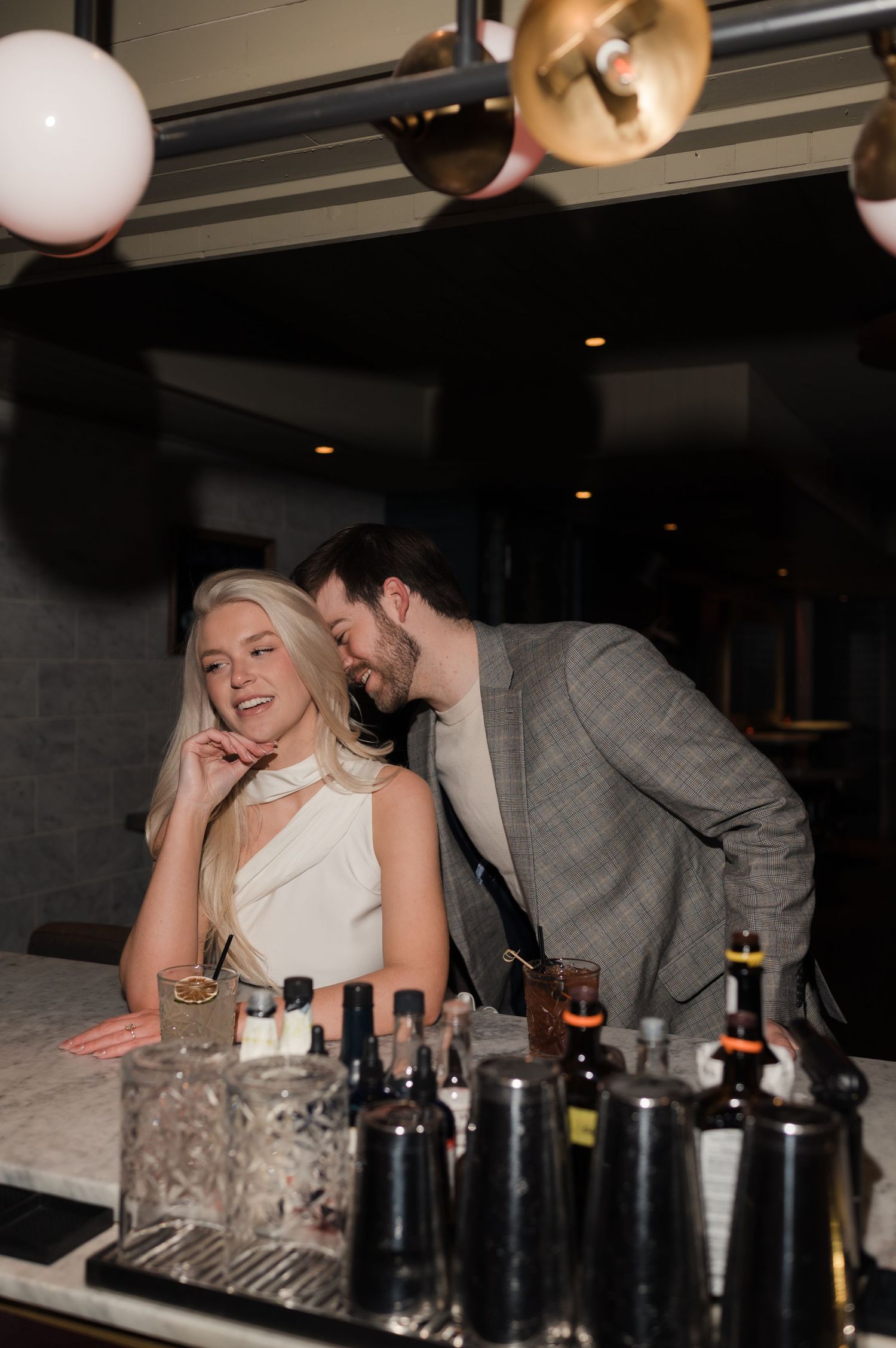 Blonde woman smiling at a bar counter as her partner whispers to her in an elegant nightlife setting.