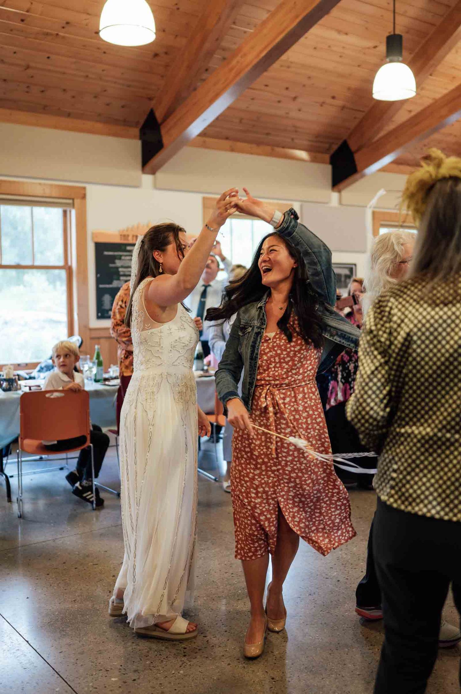 Bride and guest joyfully dancing together at a rustic indoor wedding reception with wooden ceiling.