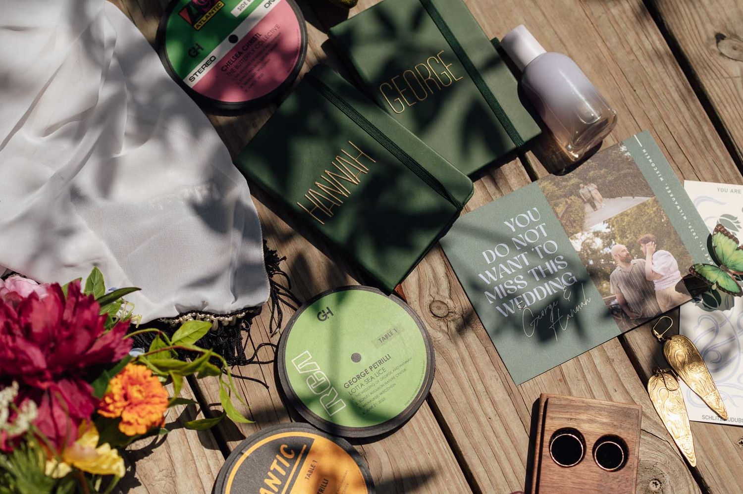 Overhead flat lay of green wedding details, candle, rings, and colorful flowers on wood.