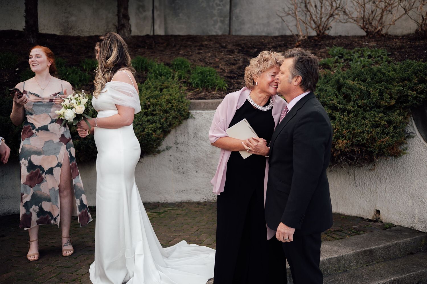 Bride in white gown greets guests at outdoor wedding ceremony with bridal party nearby.