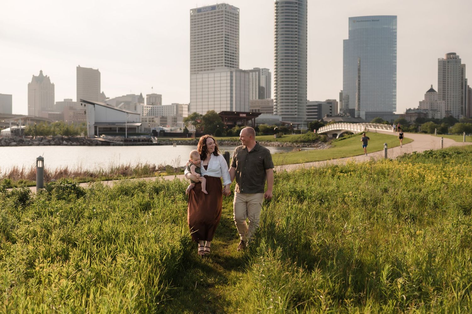 Young couple strolling through green meadow with baby, city skyline visible in the background.