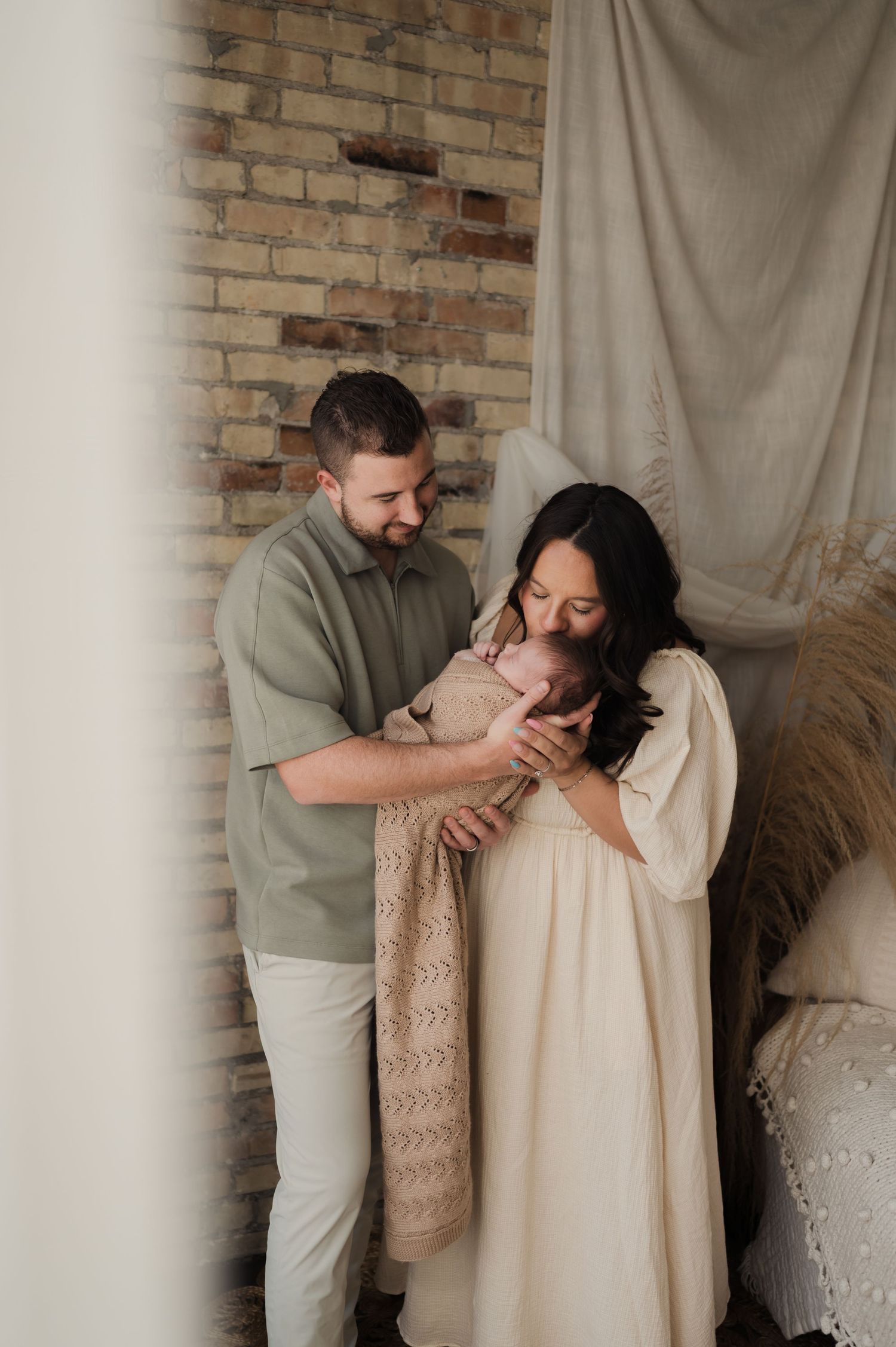 Smiling parents in neutral tones tenderly hold their newborn baby in a warm, boho-styled studio.