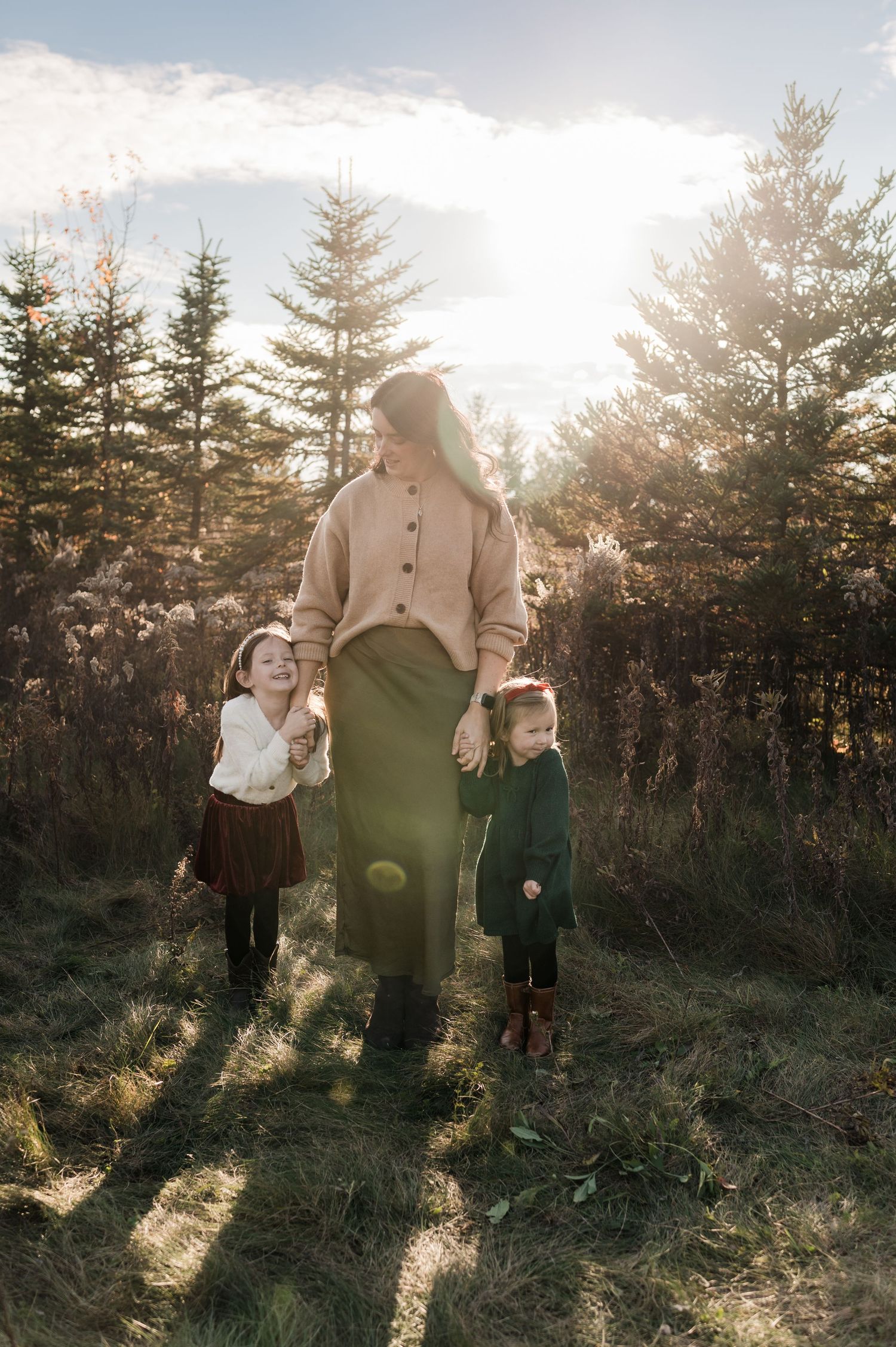Mother walking hand-in-hand with two young daughters through a sunlit pine tree farm.