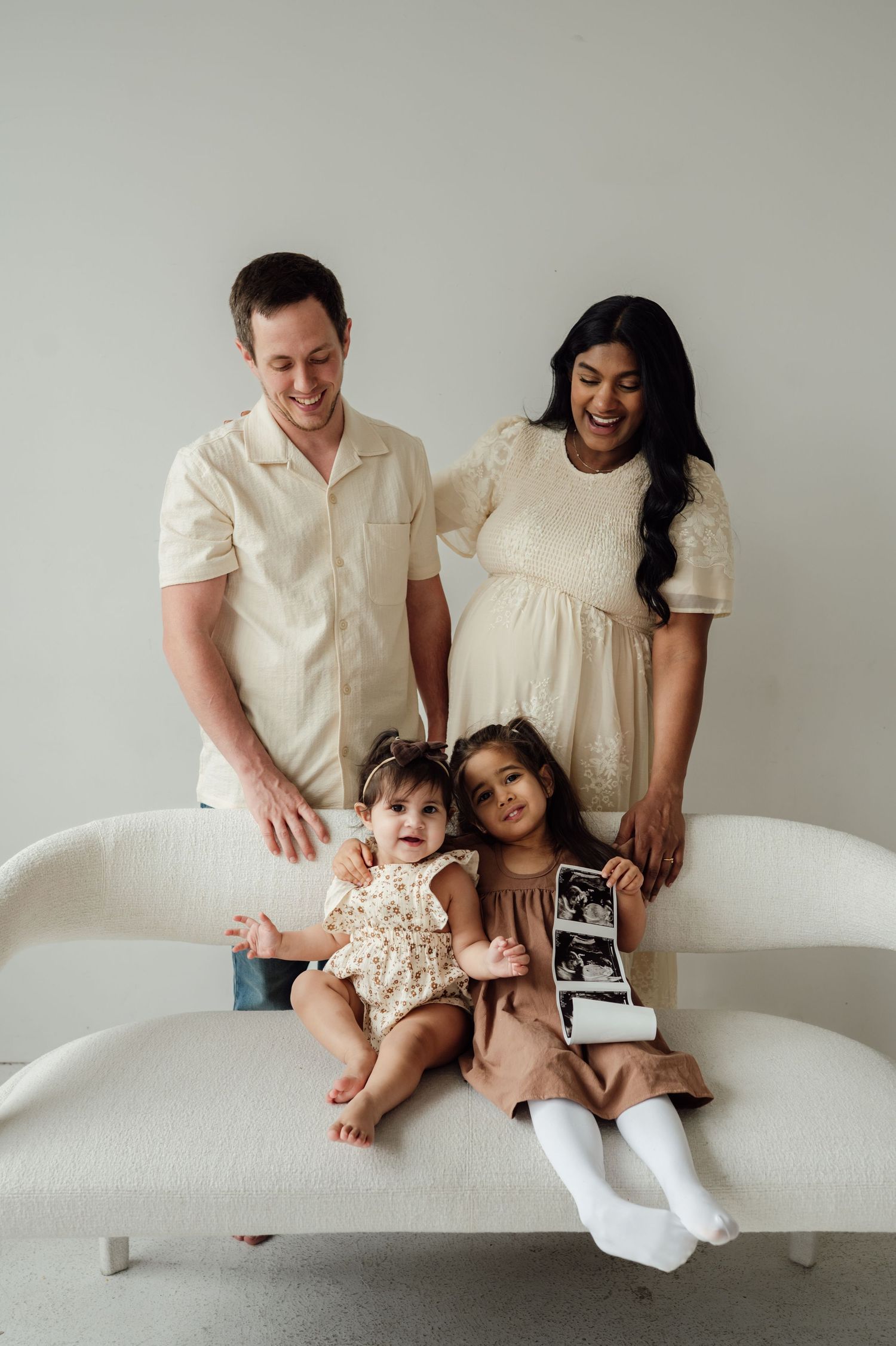 Smiling family of four with pregnant mom and two daughters holding ultrasound photos on white sofa.