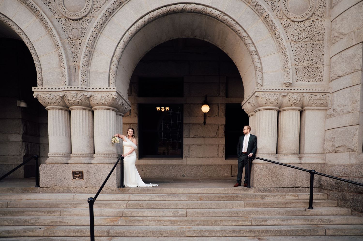 Bride and groom posing on ornate stone steps of a grand arched building entrance on their wedding day.