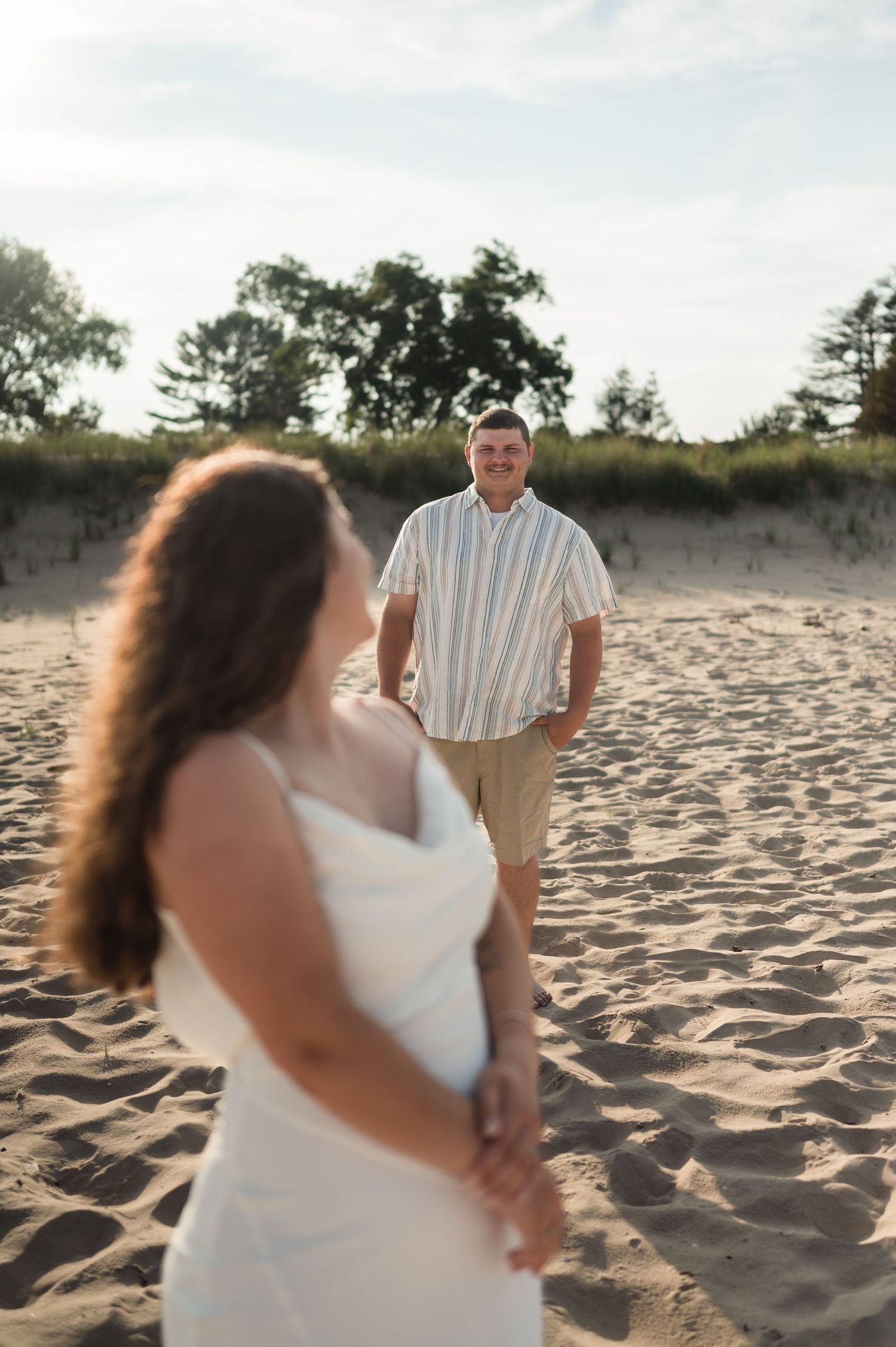 A couple shares a romantic moment on a sandy beach at sunset with sand dunes and trees in the background.