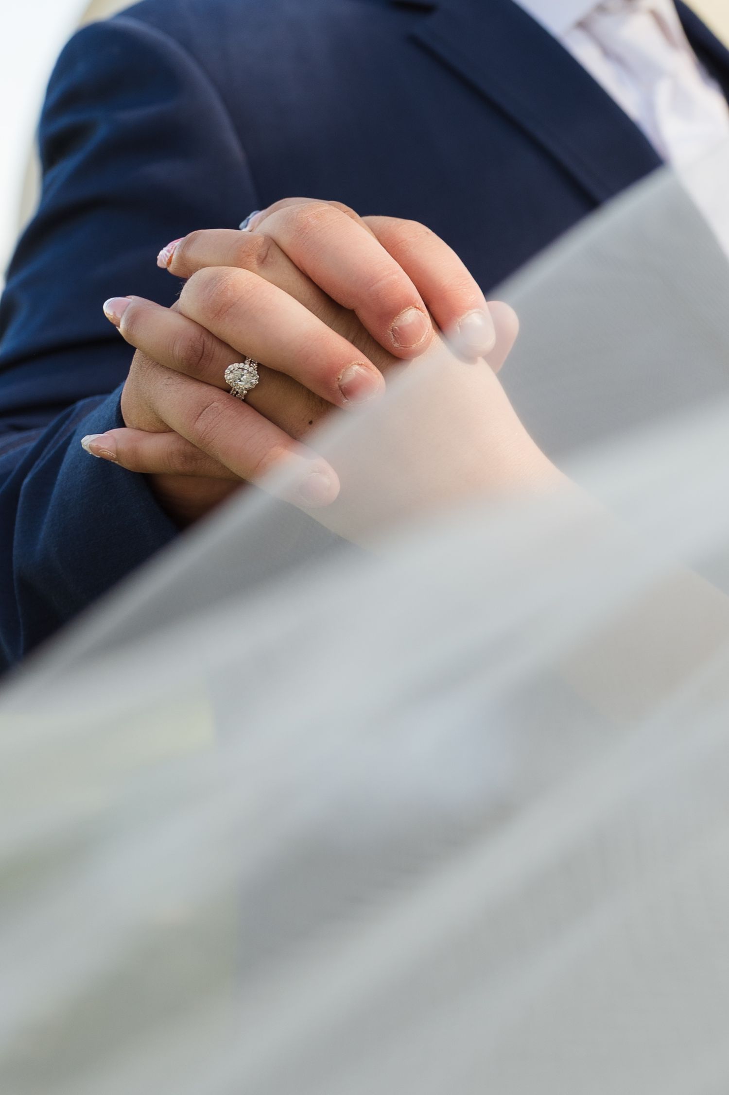 Close-up view of a sparkly diamond engagement ring on a hand with a navy blue suit sleeve in the background.