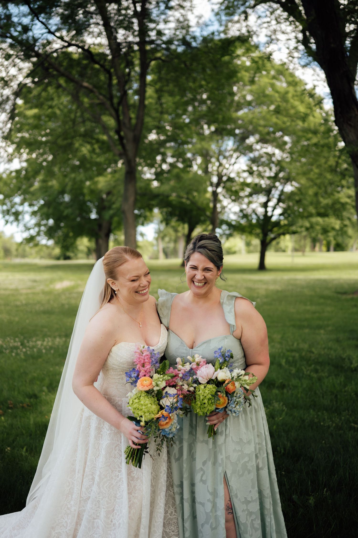 Bride in lace gown and bridesmaid in sage green dress laughing together outdoors holding colorful bouquets.