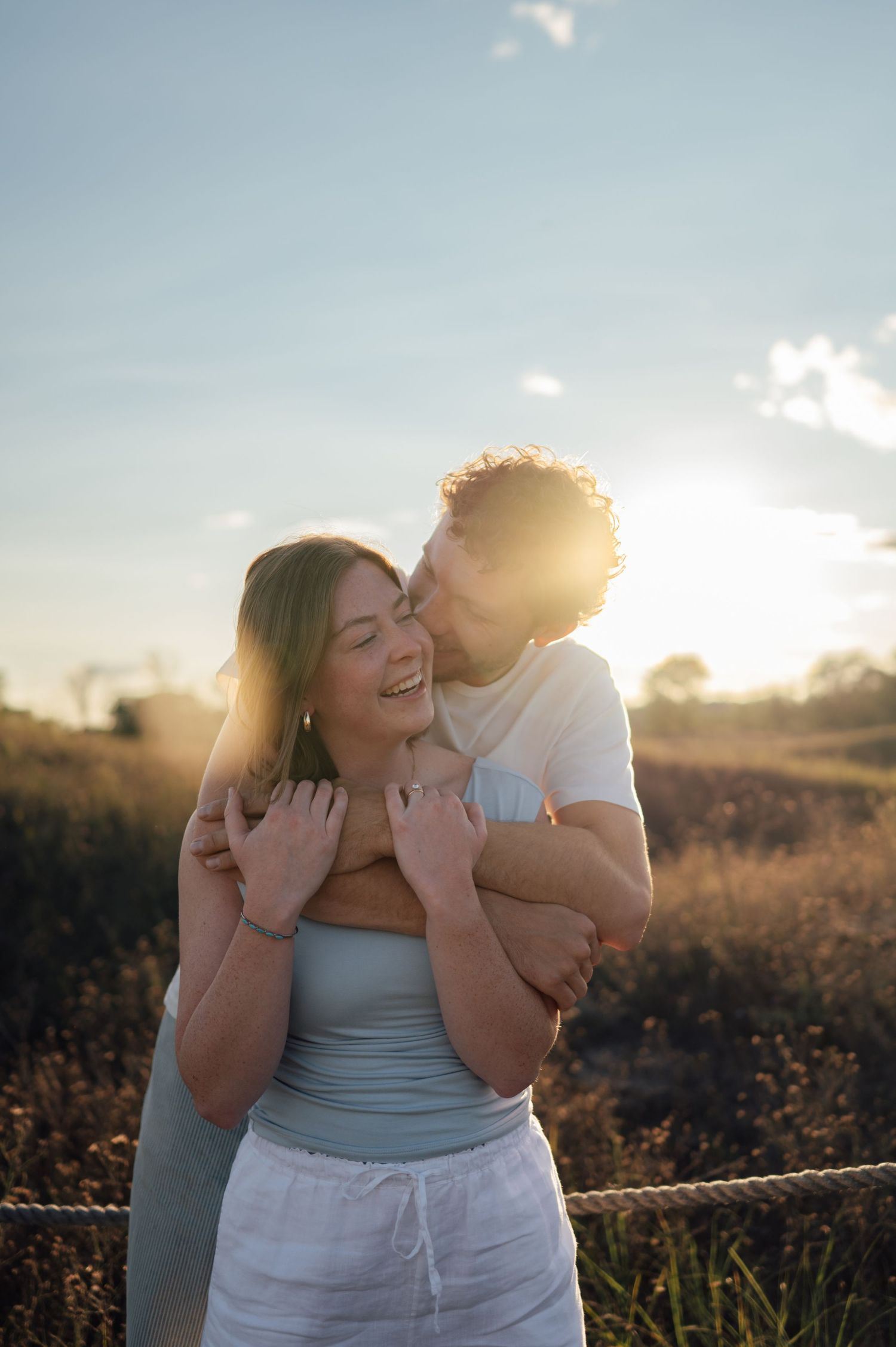 Couple embracing at golden hour in a field, man kissing woman on cheek, backlit by warm sunset glow.