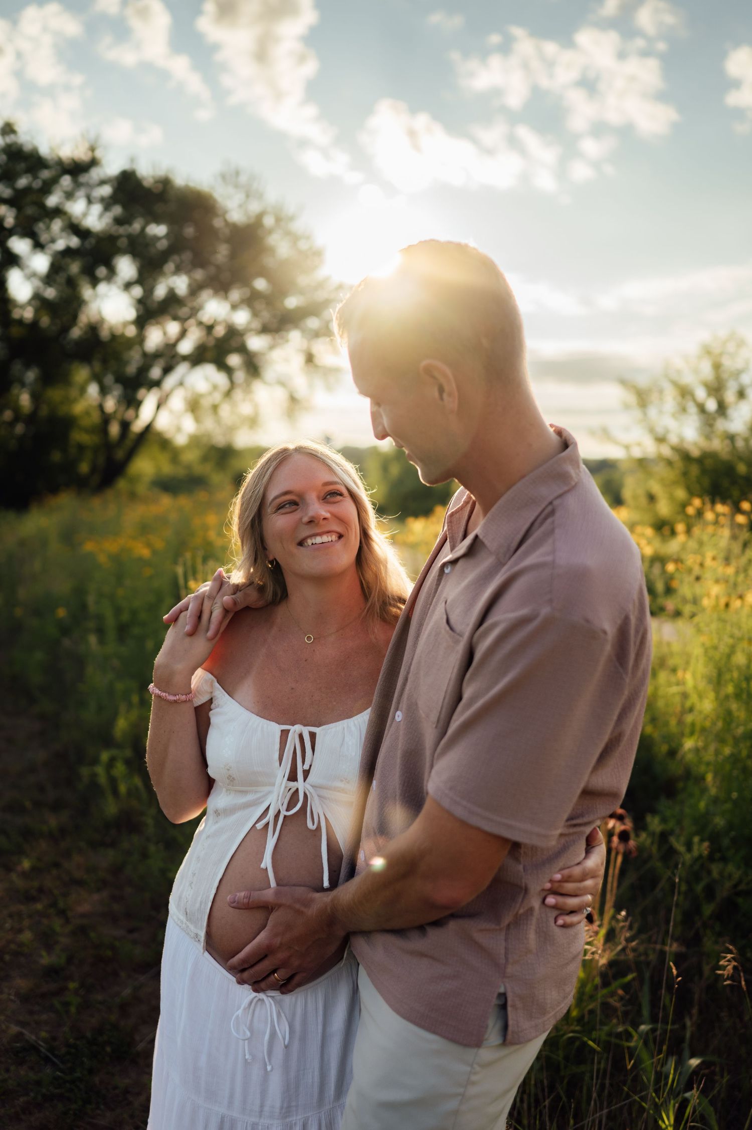 Pregnant woman in white dress smiles at partner during golden hour maternity photoshoot in a lush green field.