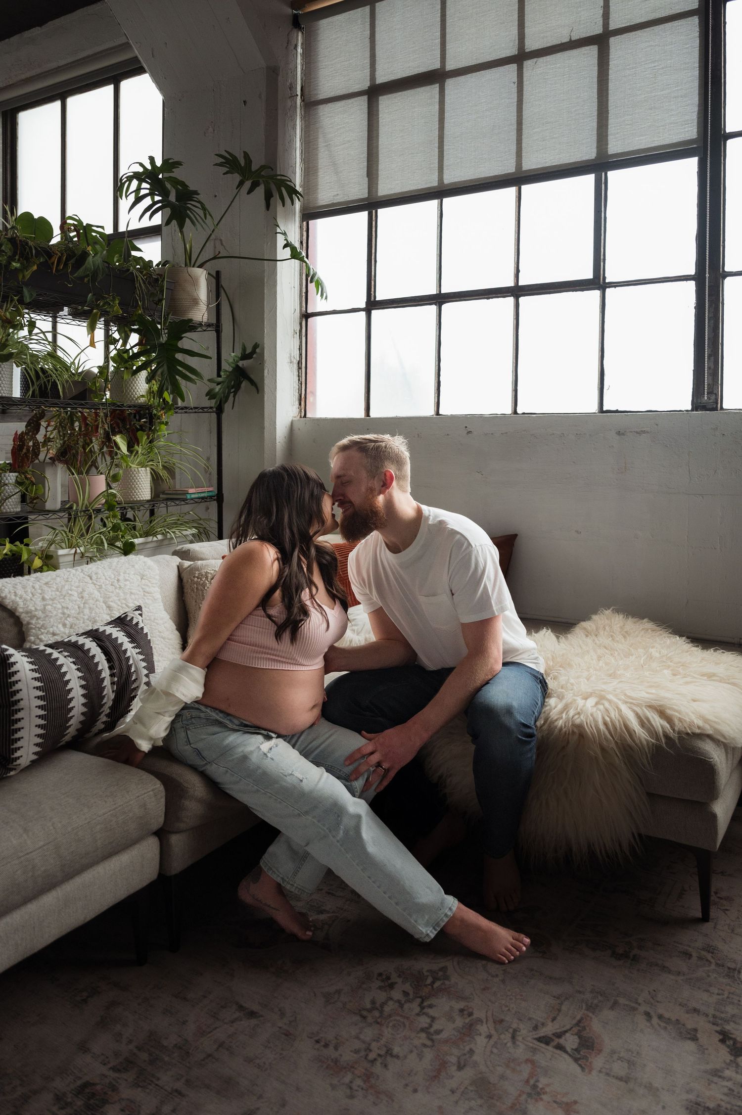 Pregnant couple sharing a tender kiss on a couch in a cozy loft apartment surrounded by plants.