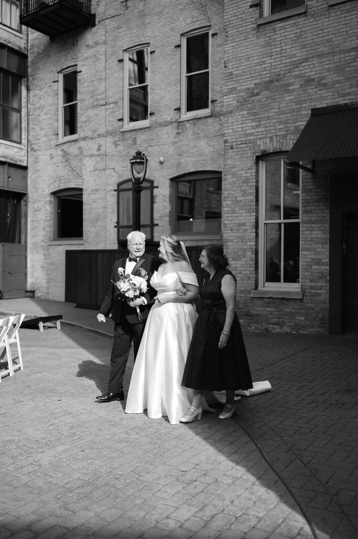 Black and white photo of bride in white gown with parents outside a historic brick building before ceremony.