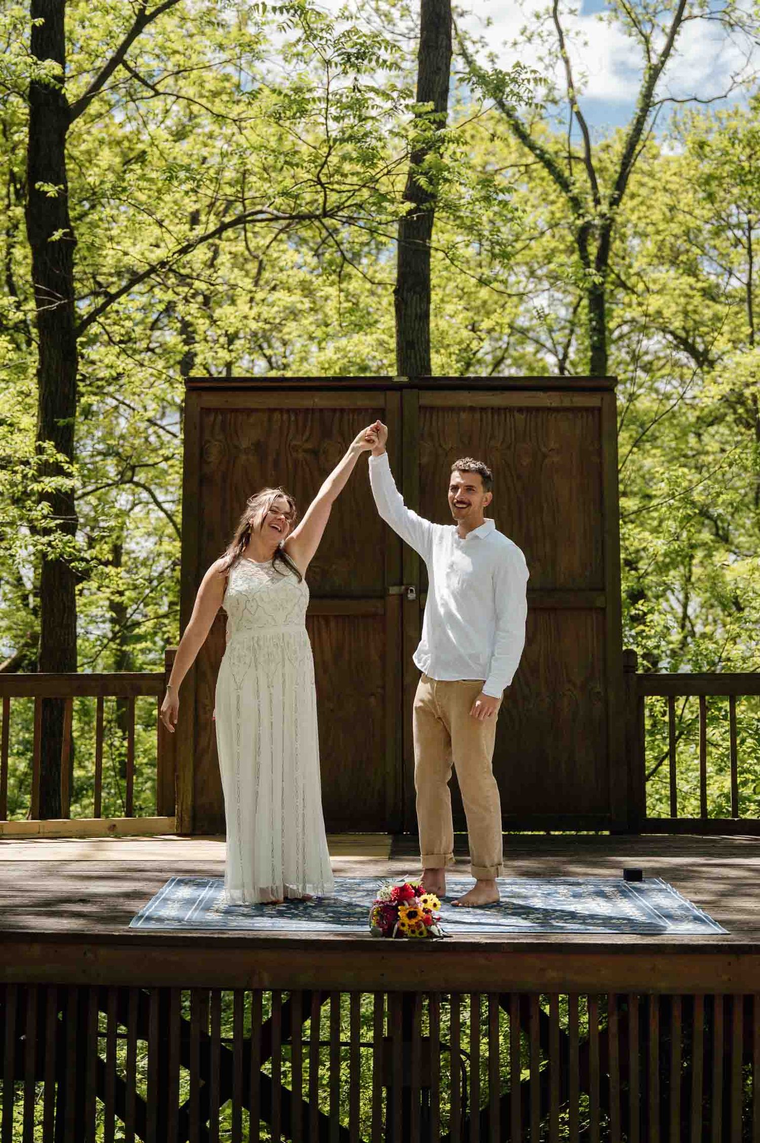 Bride and groom celebrating on wooden deck surrounded by lush green trees after ceremony.