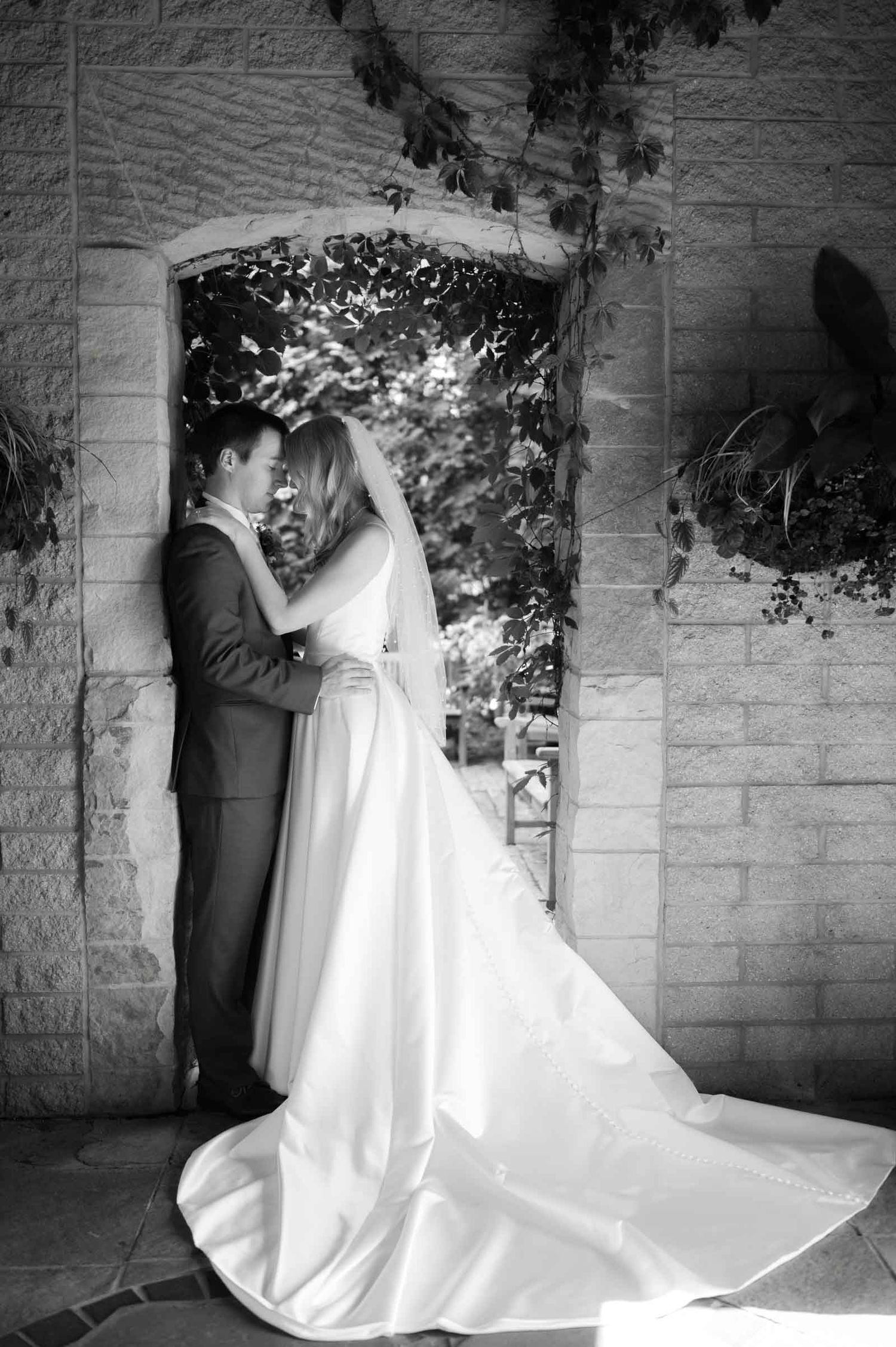 Black and white romantic portrait of bride and groom embracing under stone archway with flowing wedding gown train.