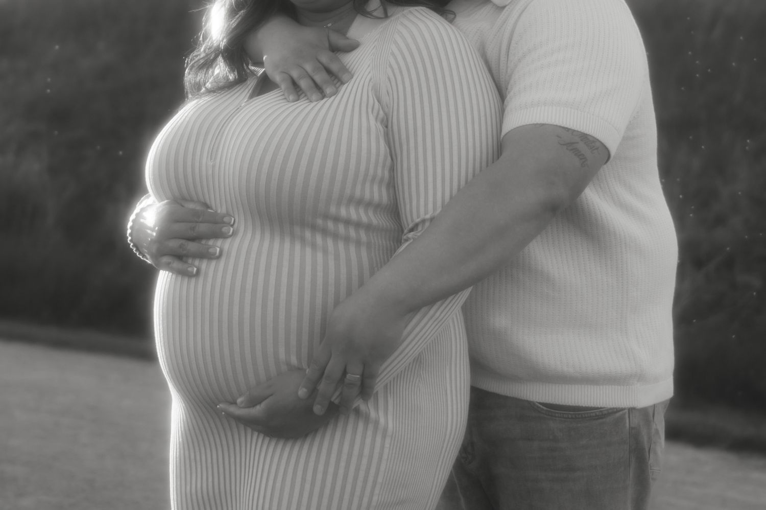 Black and white photo of pregnant couple cradling baby bump outdoors.