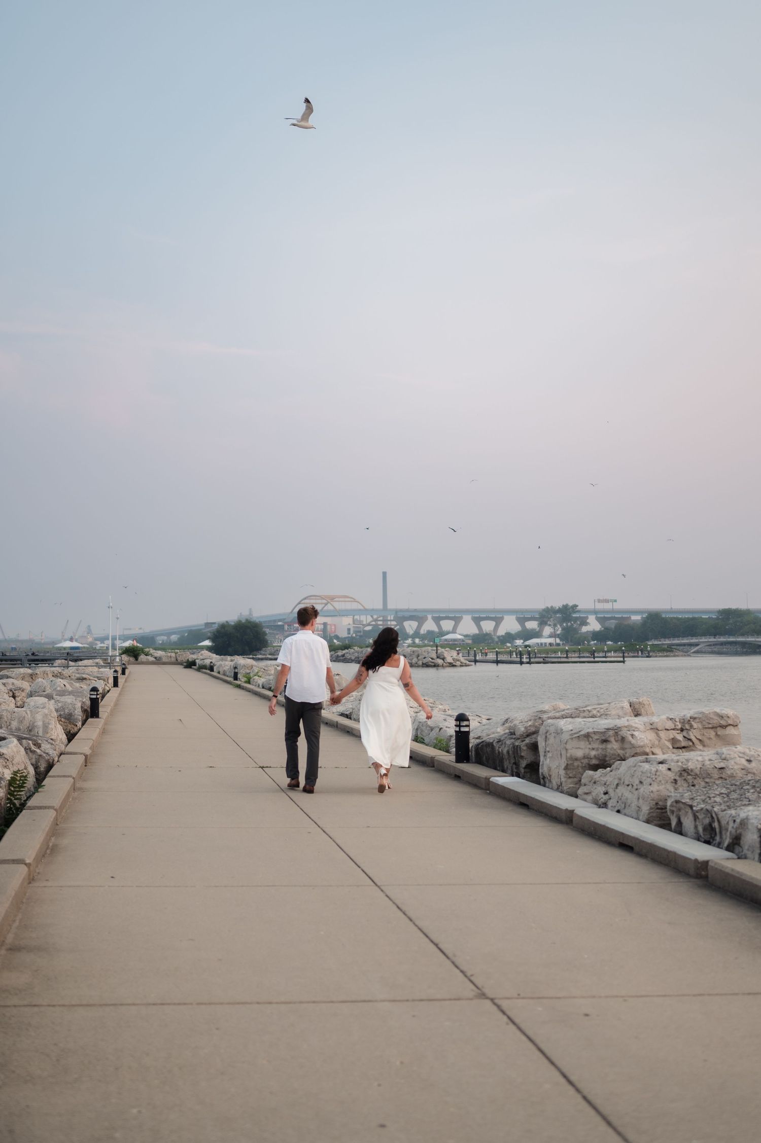 Engaged couple walking hand-in-hand along a lakeside pier with city bridge in background.