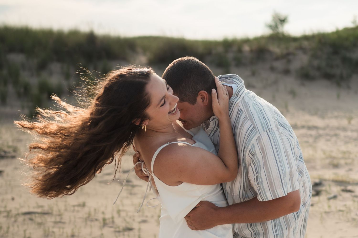 A romantic couple embraces and kisses on a beach boardwalk at sunset, with long hair flowing in the wind.