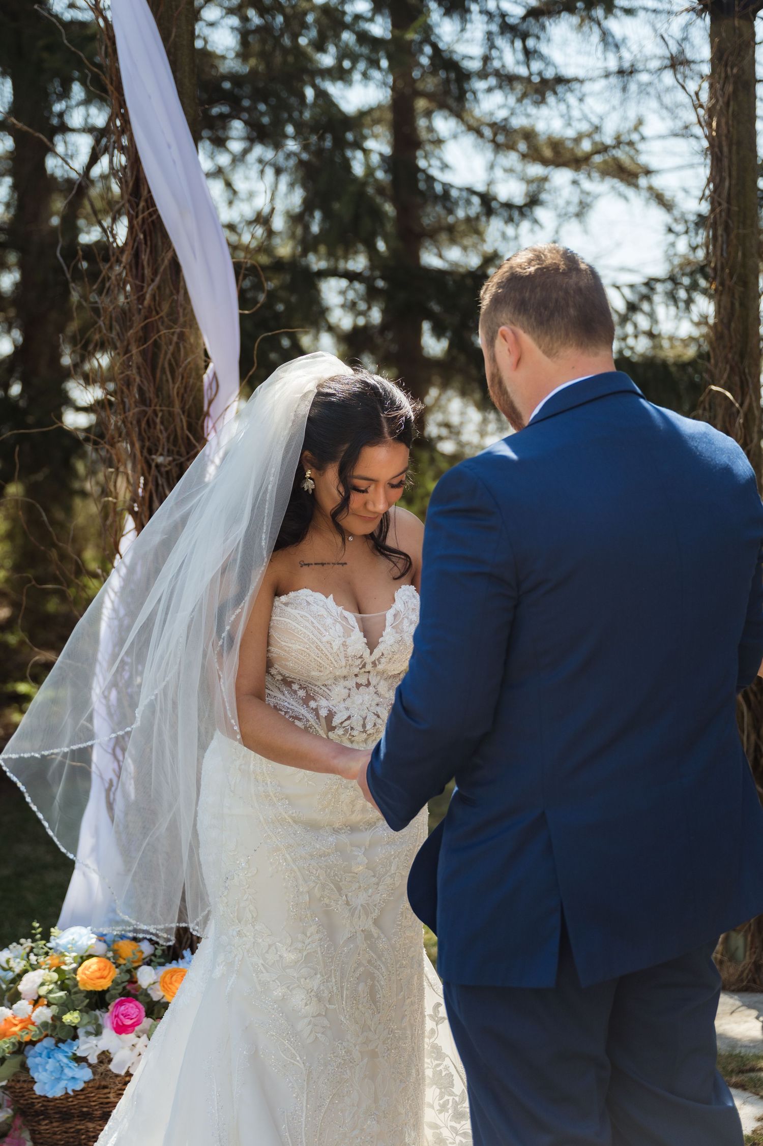 Wedding couple at outdoor forest ceremony with bride in white lace dress and groom in navy suit standing by floral decor.