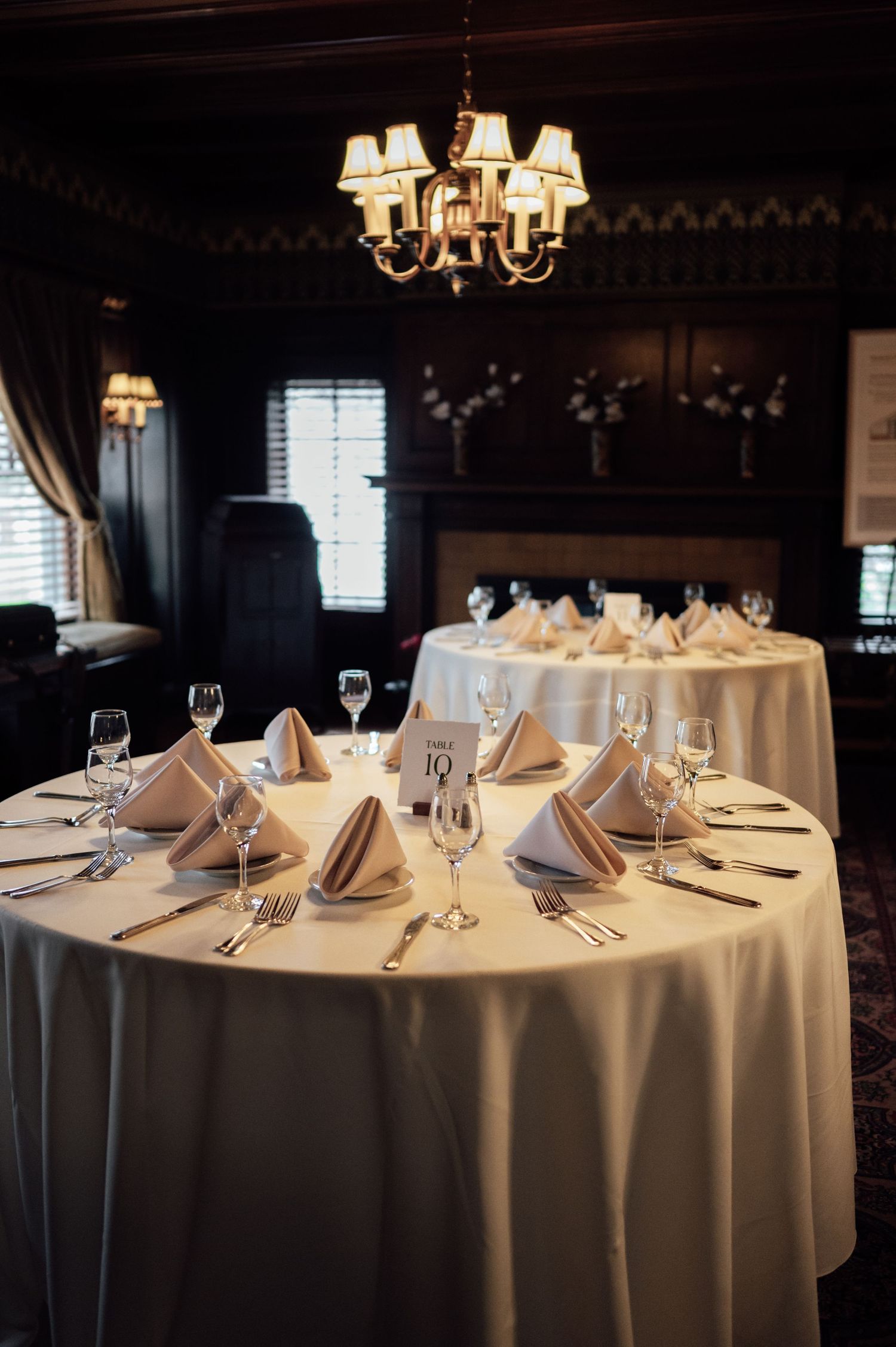 Elegant banquet room with round tables set with white linens, folded napkins, crystal glasses, and a chandelier overhead.
