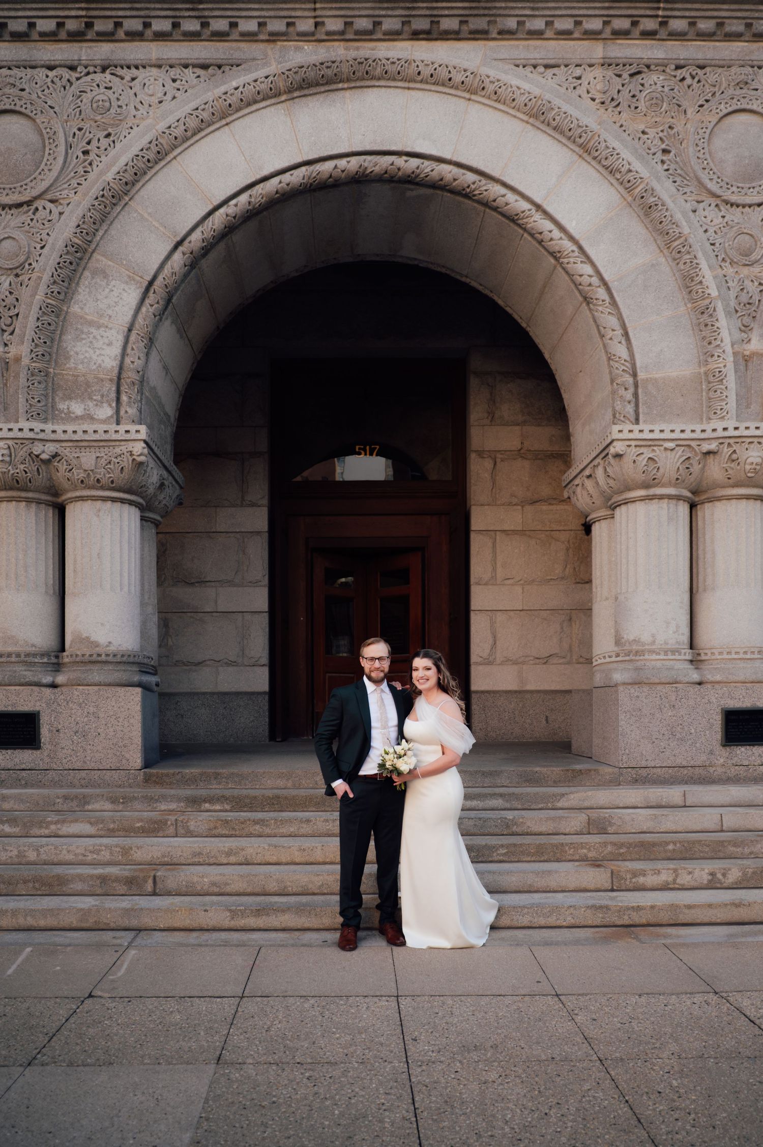 Bride and groom posing under ornate stone arch at elegant building entrance on their wedding day.