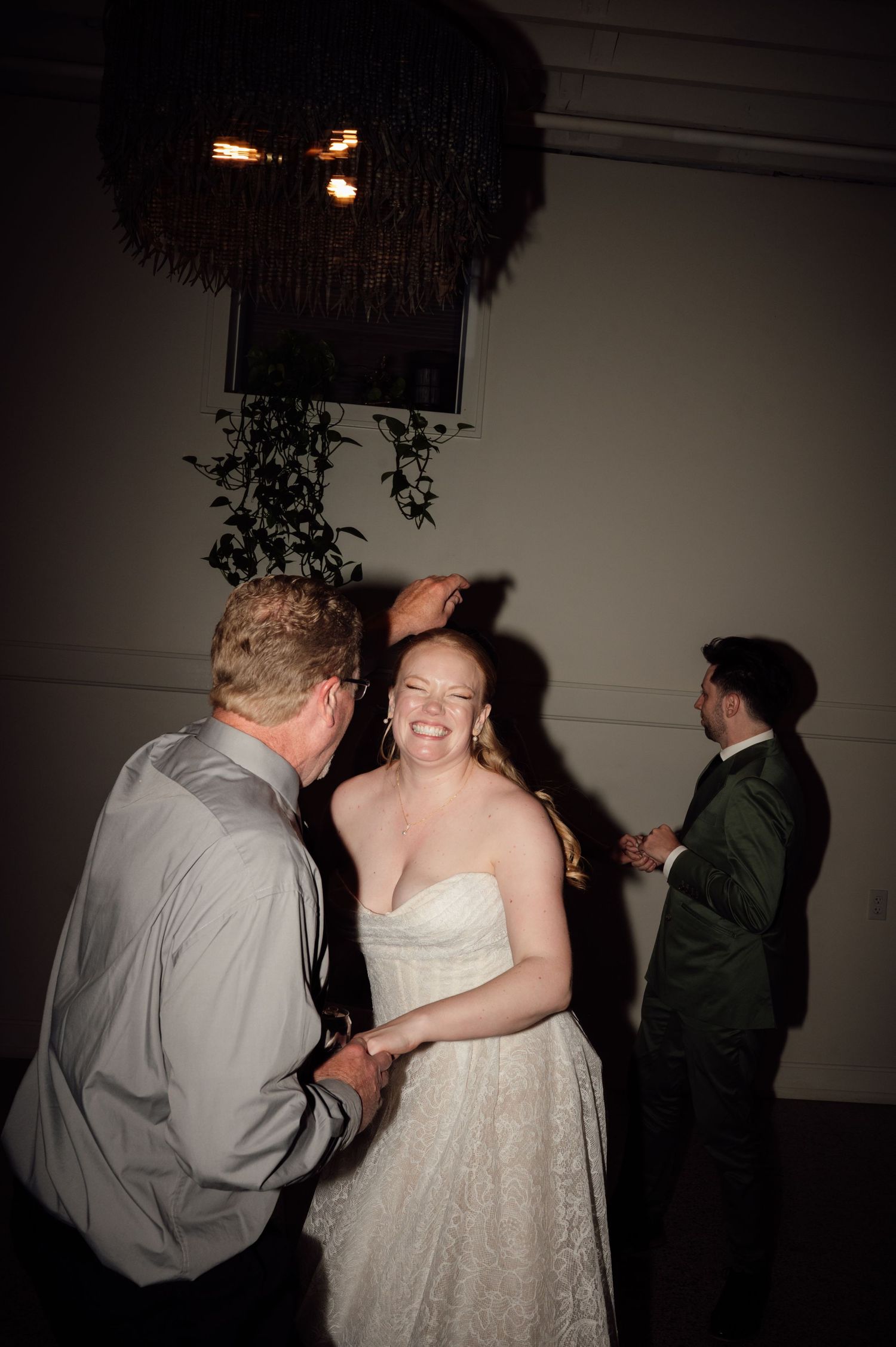 Bride in strapless lace gown laughing while dancing with a man in gray suit at a dimly lit wedding reception.