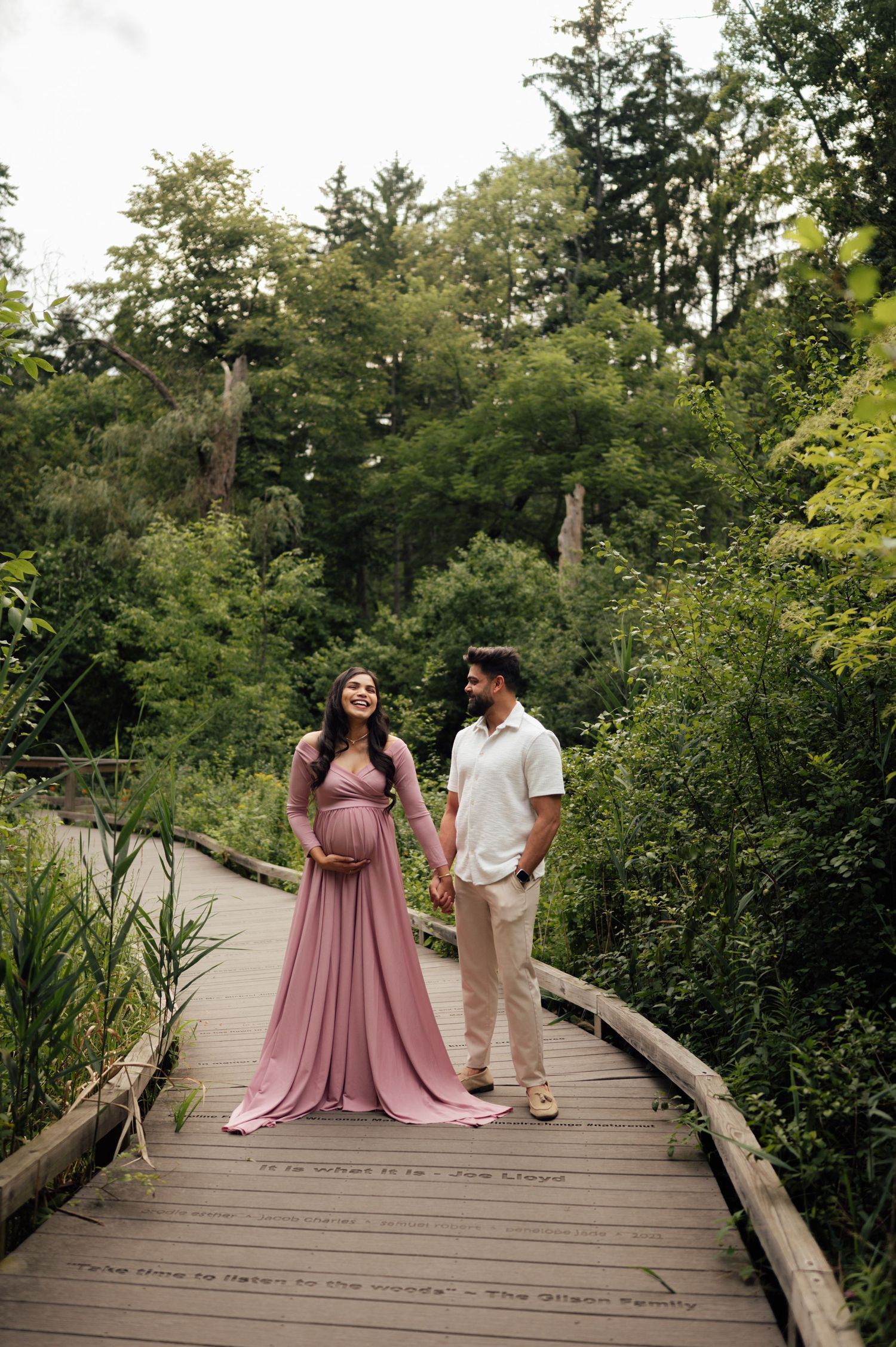 Pregnant woman in pink maternity gown holds hands with partner on wooden boardwalk surrounded by lush greenery.