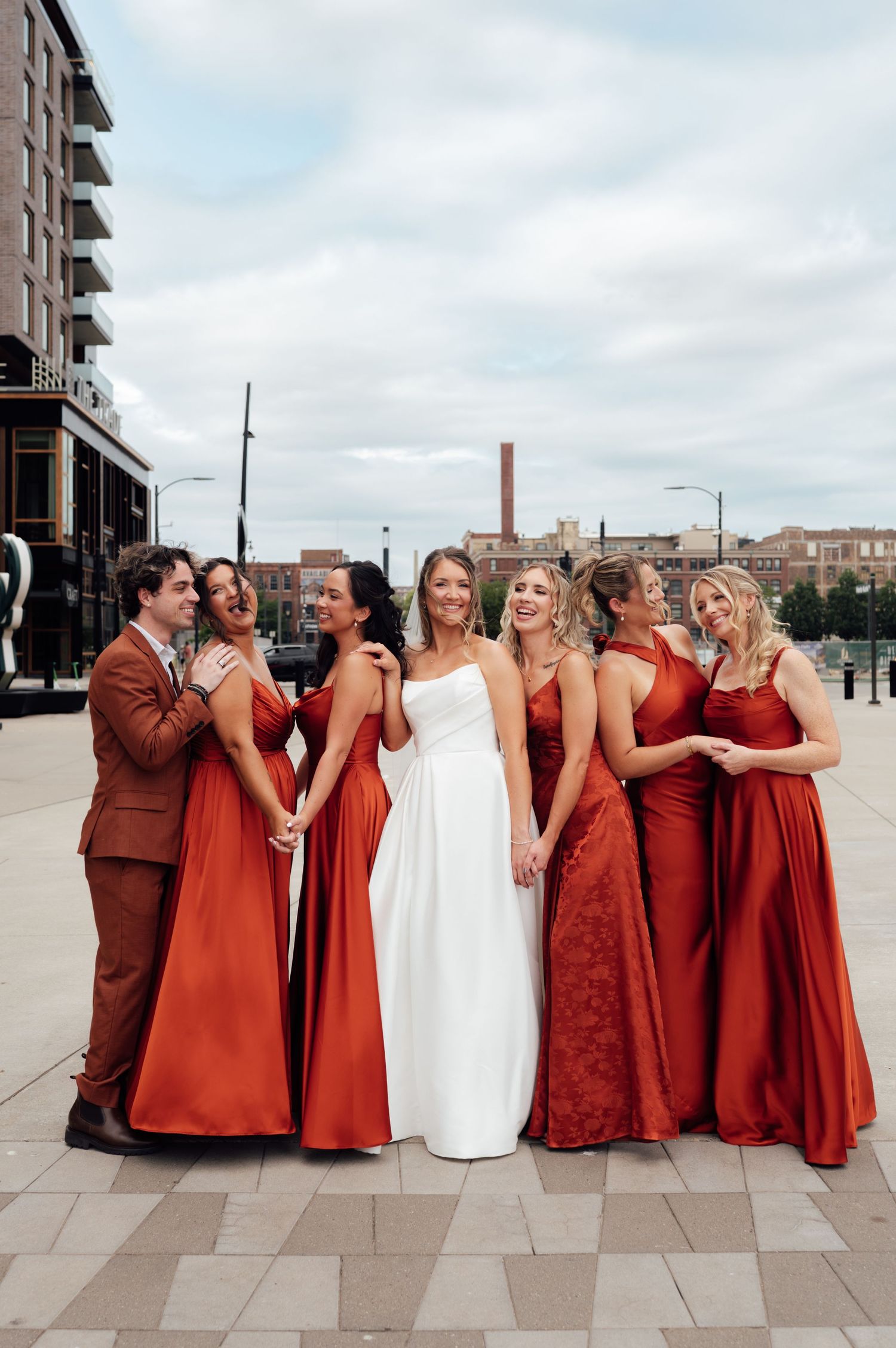 Bride in white gown poses with bridesmaids in rust-red dresses outdoors at an urban waterfront venue.