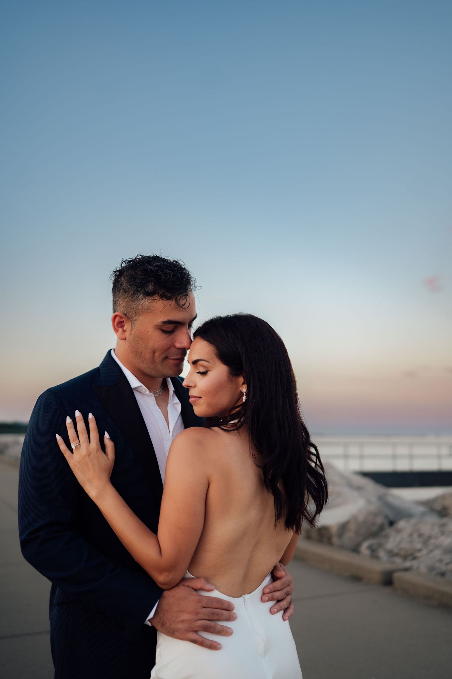 Couple embracing at sunset, bride in backless white gown and groom in navy suit, near waterfront.