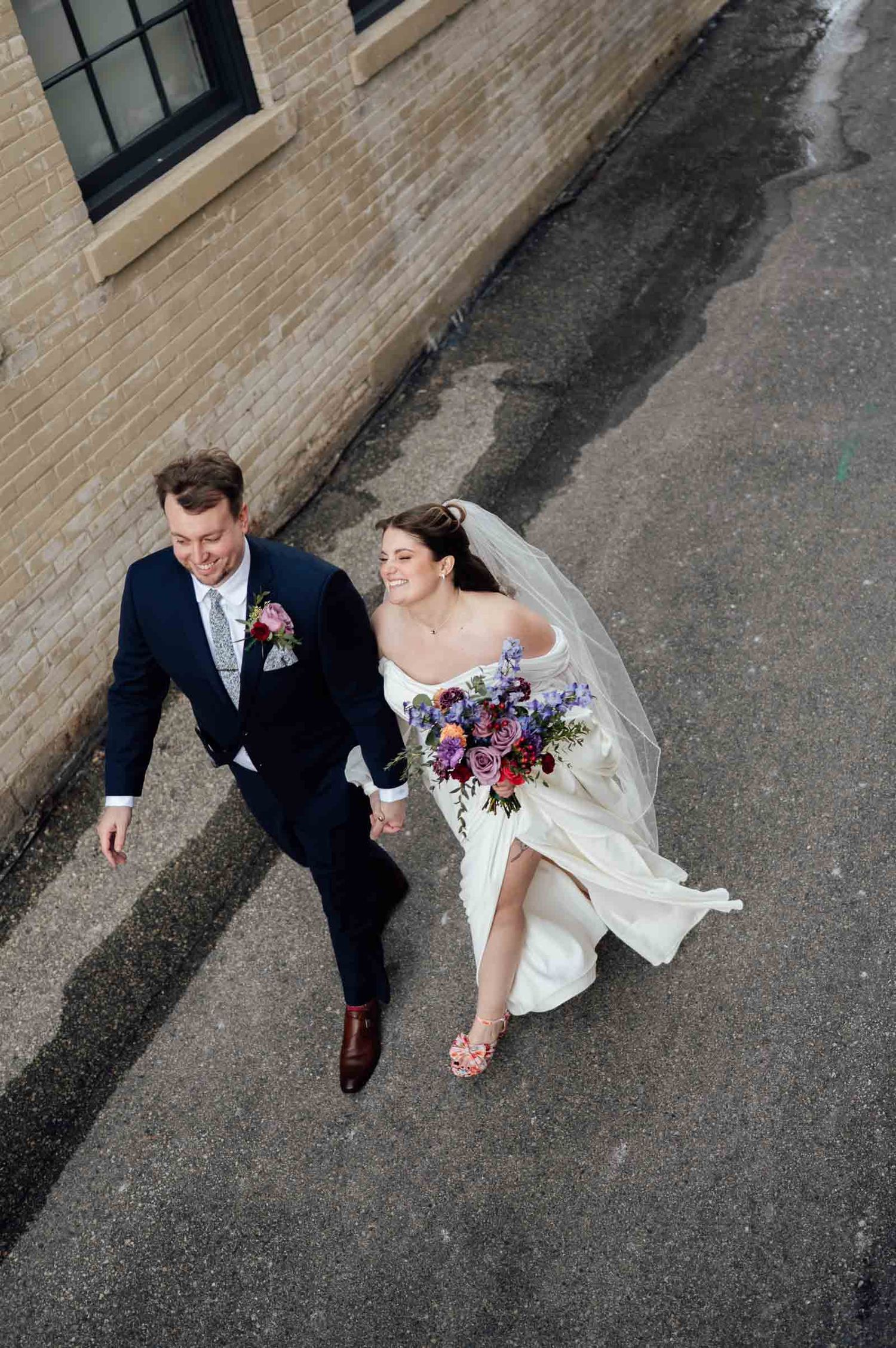 Aerial view of bride and groom walking hand in hand on wet pavement, bride holding colorful bouquet in white gown.
