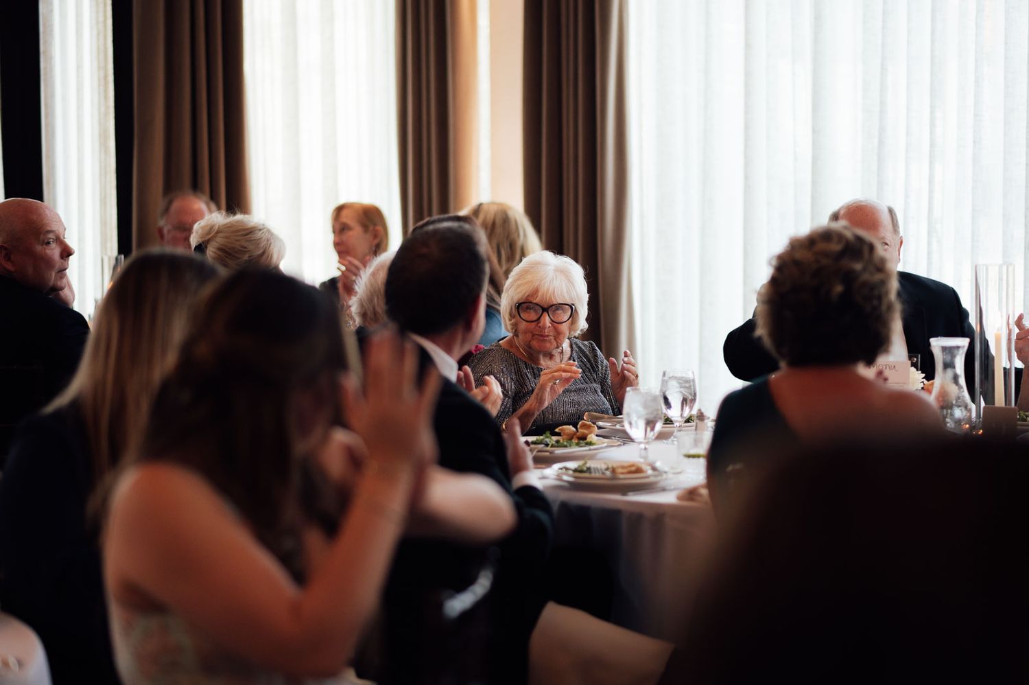Wedding guests seated at round tables enjoying dinner conversation in a warmly lit reception venue.