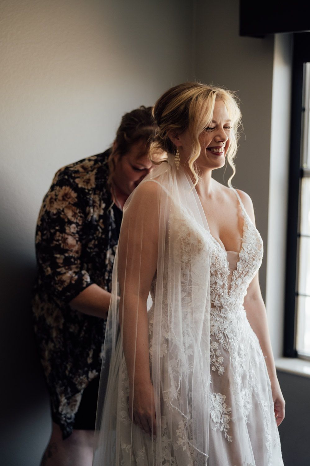 Bridesmaid helping laughing blonde bride fasten lace wedding dress near window before ceremony.