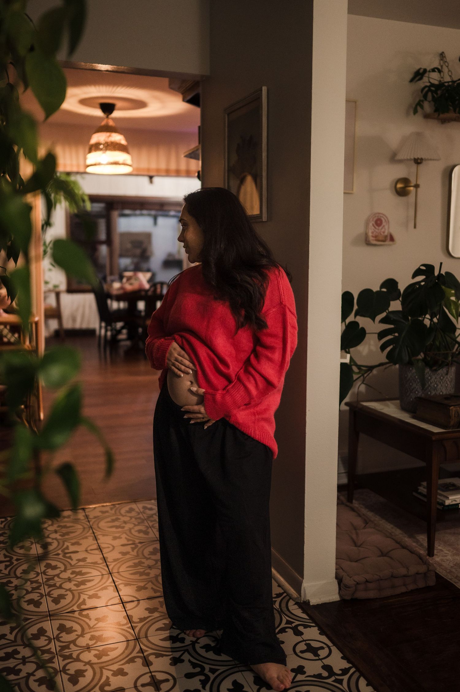 Pregnant woman in red sweater and black pants cradling baby bump in warmly lit home hallway.