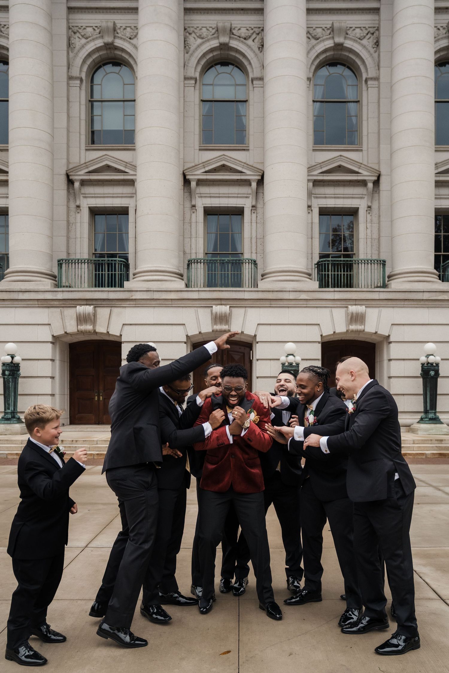 Groomsmen perform a fun choreographed leaning pose in front of a grand building with columns.