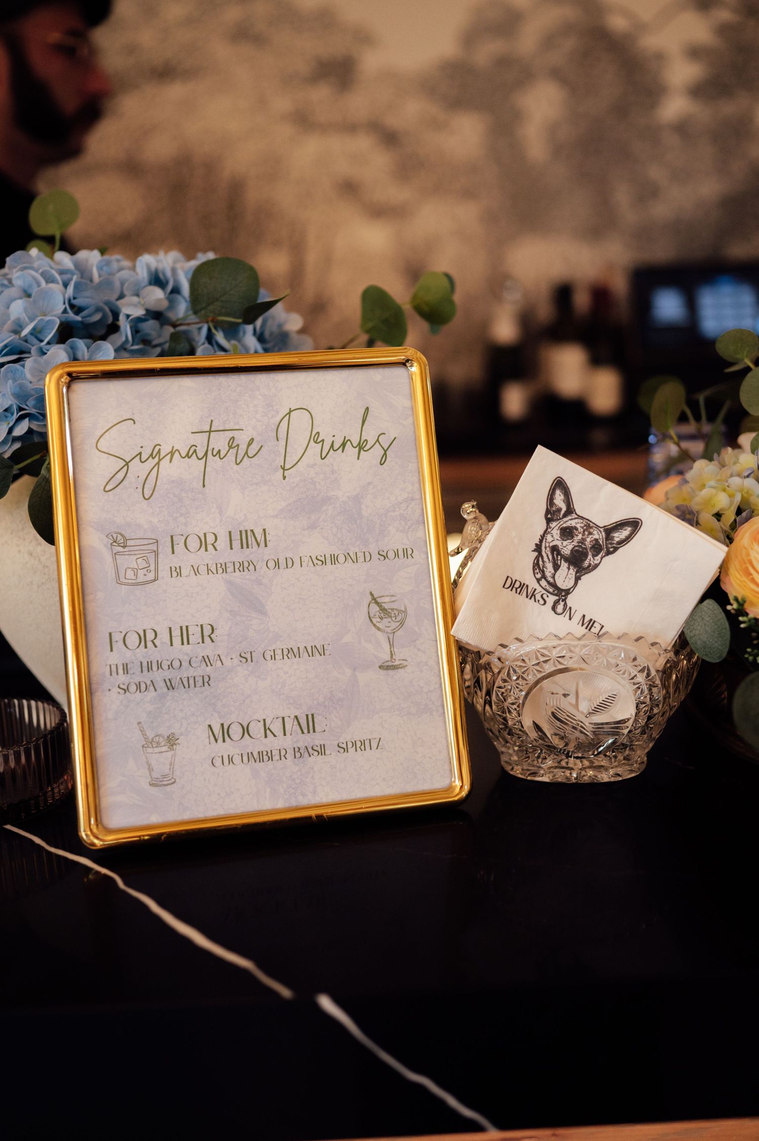Gold-framed signature drinks menu at wedding bar display with floral arrangements and dog-illustrated cocktail napkins.