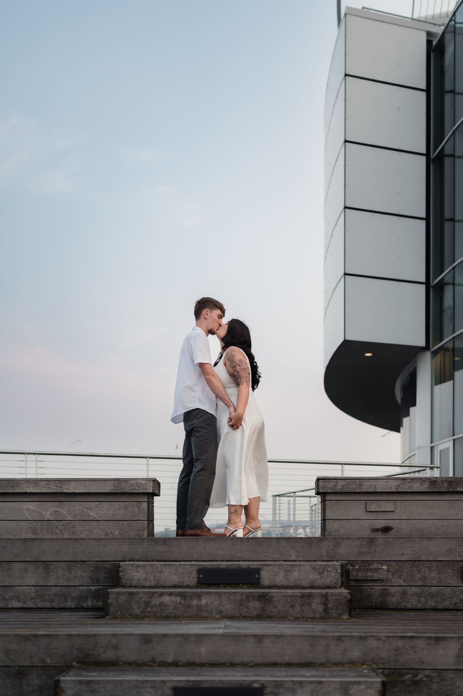 A couple shares a romantic moment on concrete steps outside a modern building at dusk.