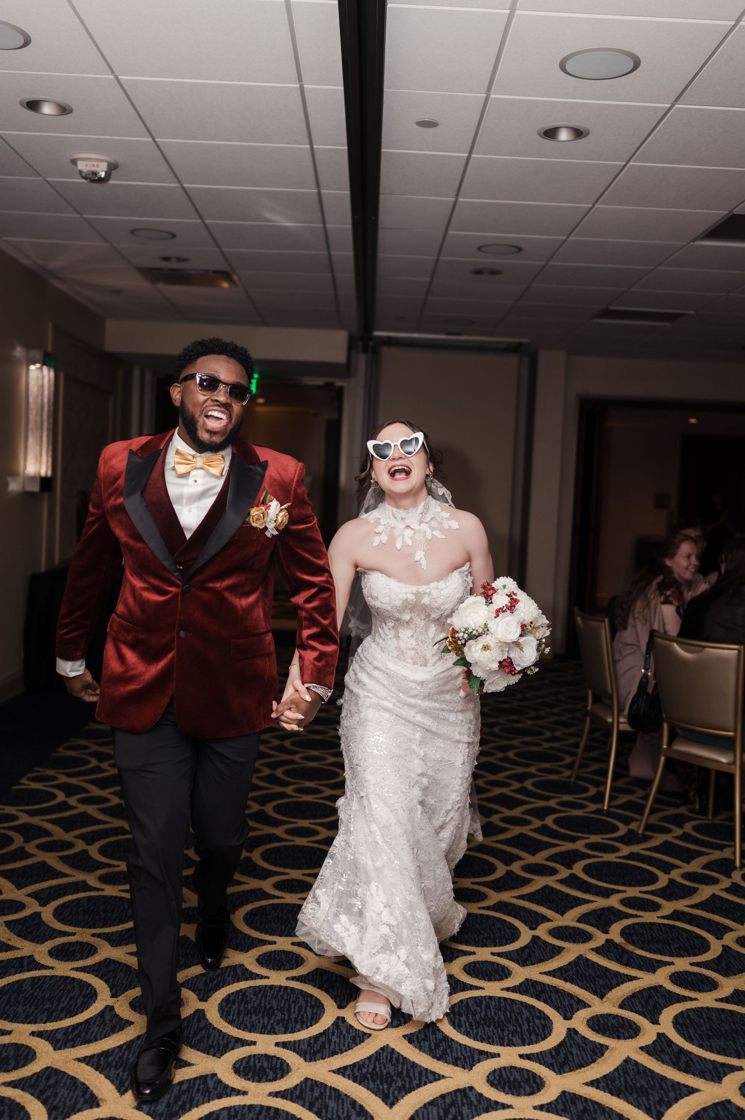 A couple wearing wedding attire and sunglasses dance together on a patterned carpet at their reception celebration.